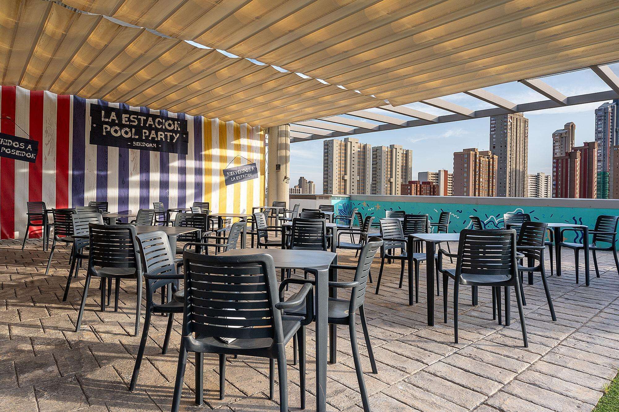 An outdoor seating area with tables and chairs, a striped wall with signs, and a cityscape view.