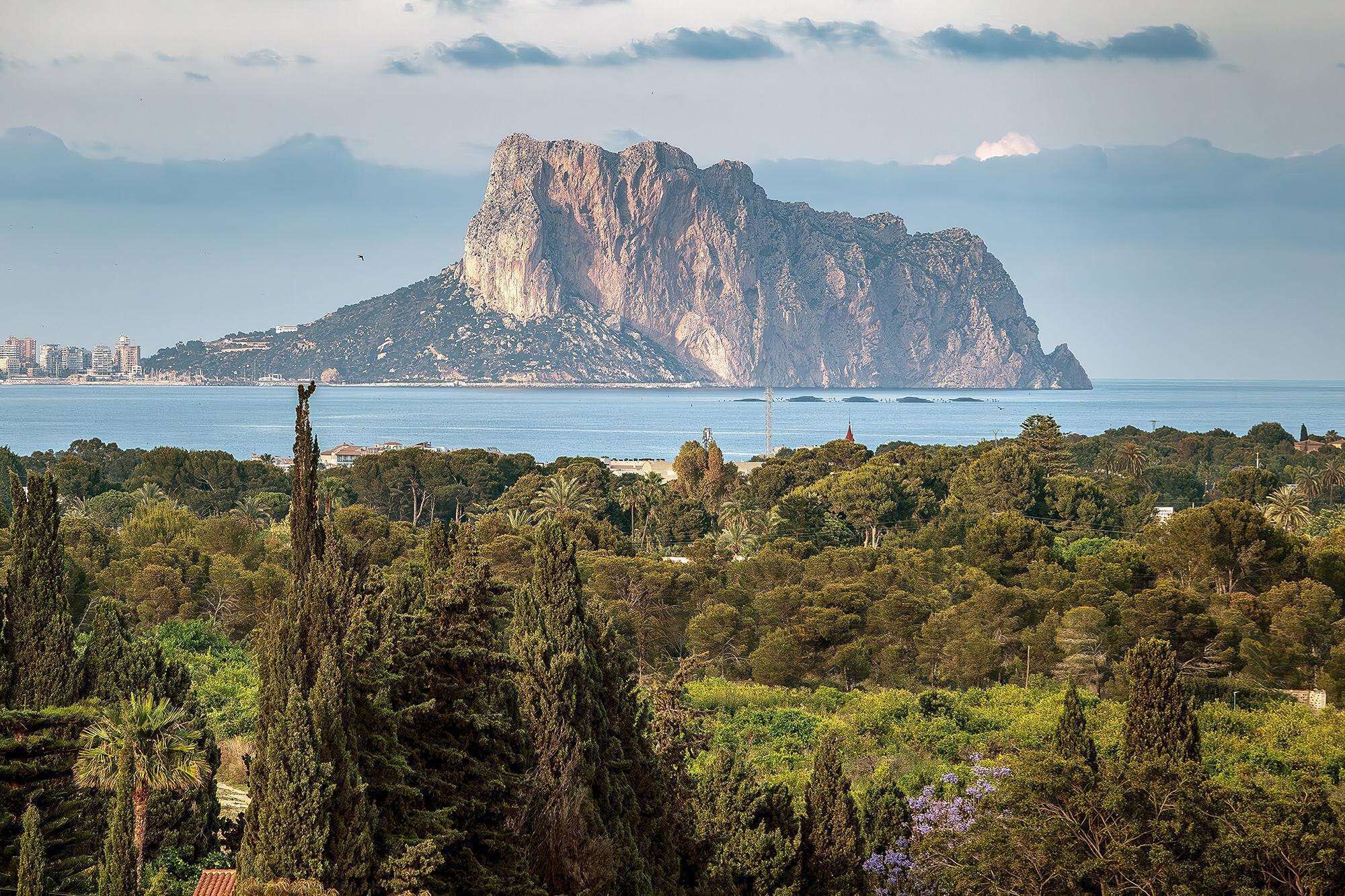 A coastal landscape with a prominent rocky mountain in the background and dense greenery in the foreground.