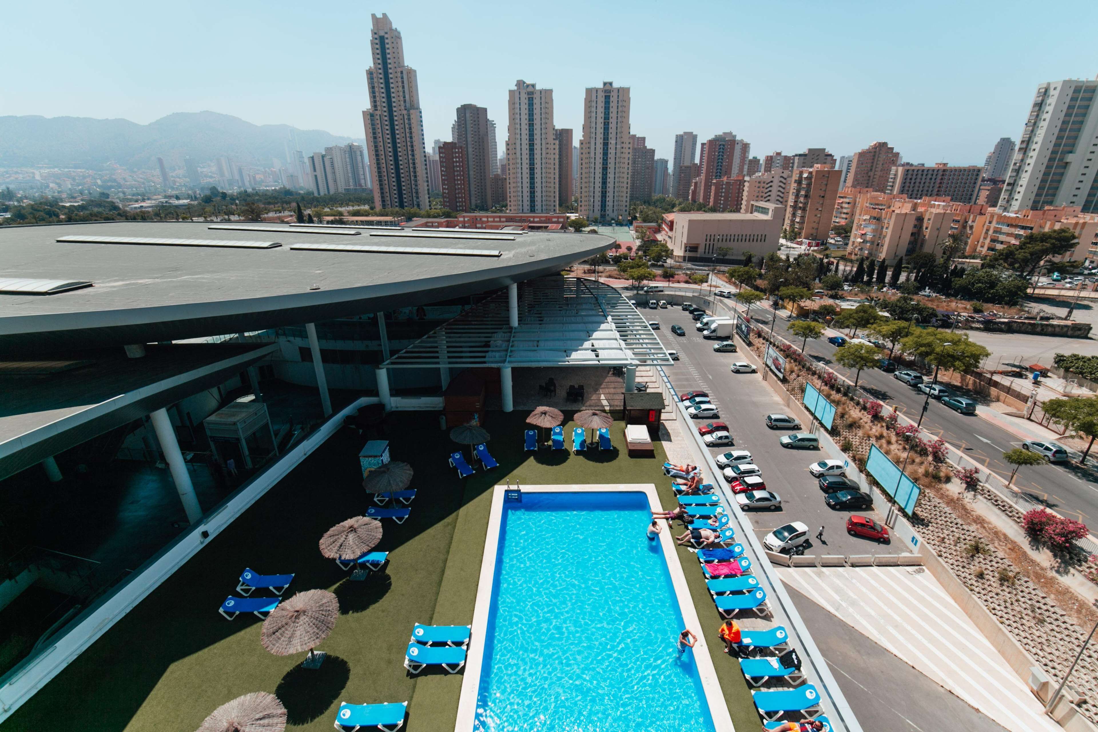A rooftop swimming pool with lounge chairs and umbrellas, surrounded by a parking area and city buildings in the background.