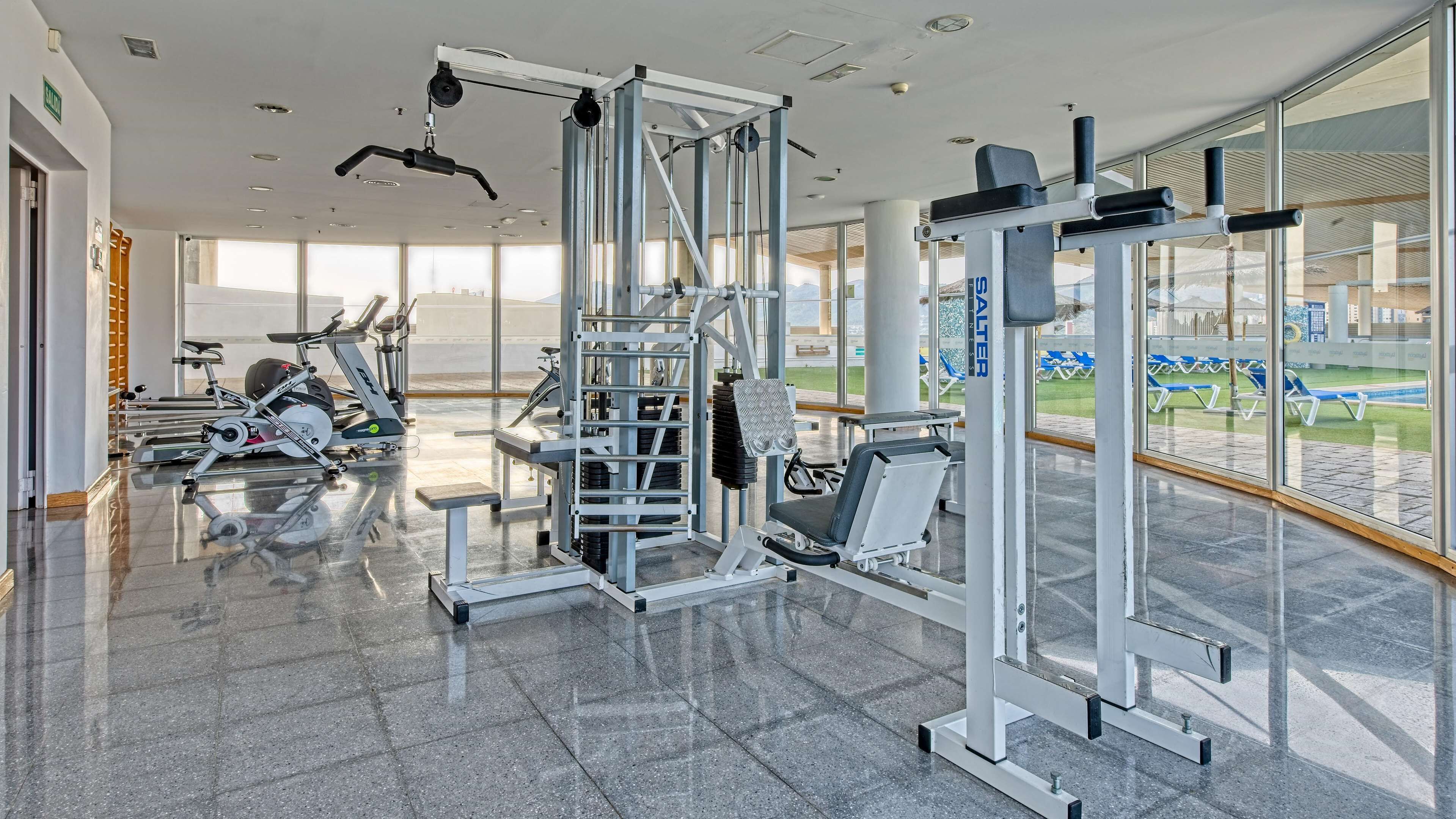 A modern gym with various exercise equipment, large windows, and a view of an outdoor pool area.