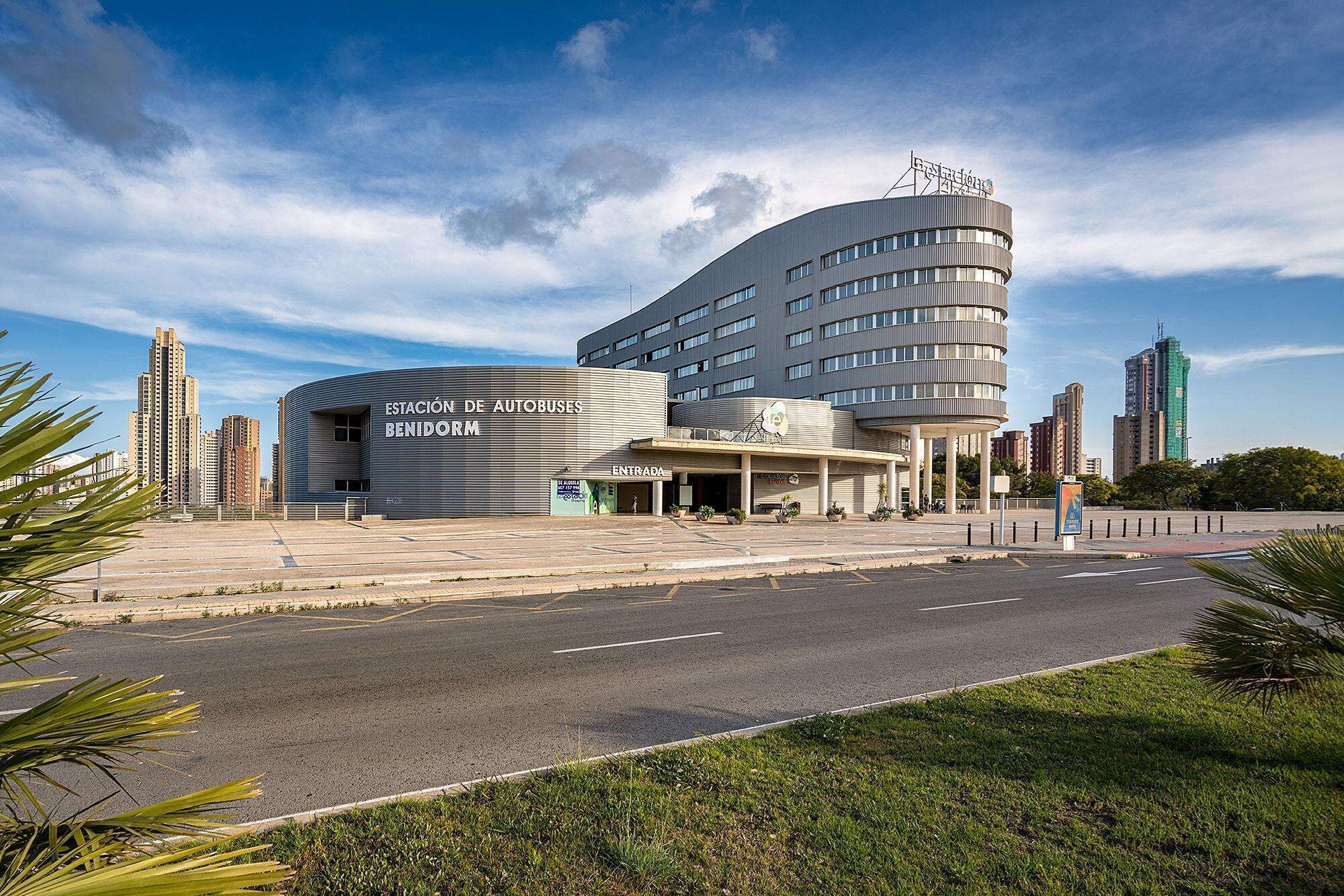 A modern bus station with a curved facade and multiple levels, featuring a prominent entrance and a sign for 'Estación de Autobuses Benidorm'.