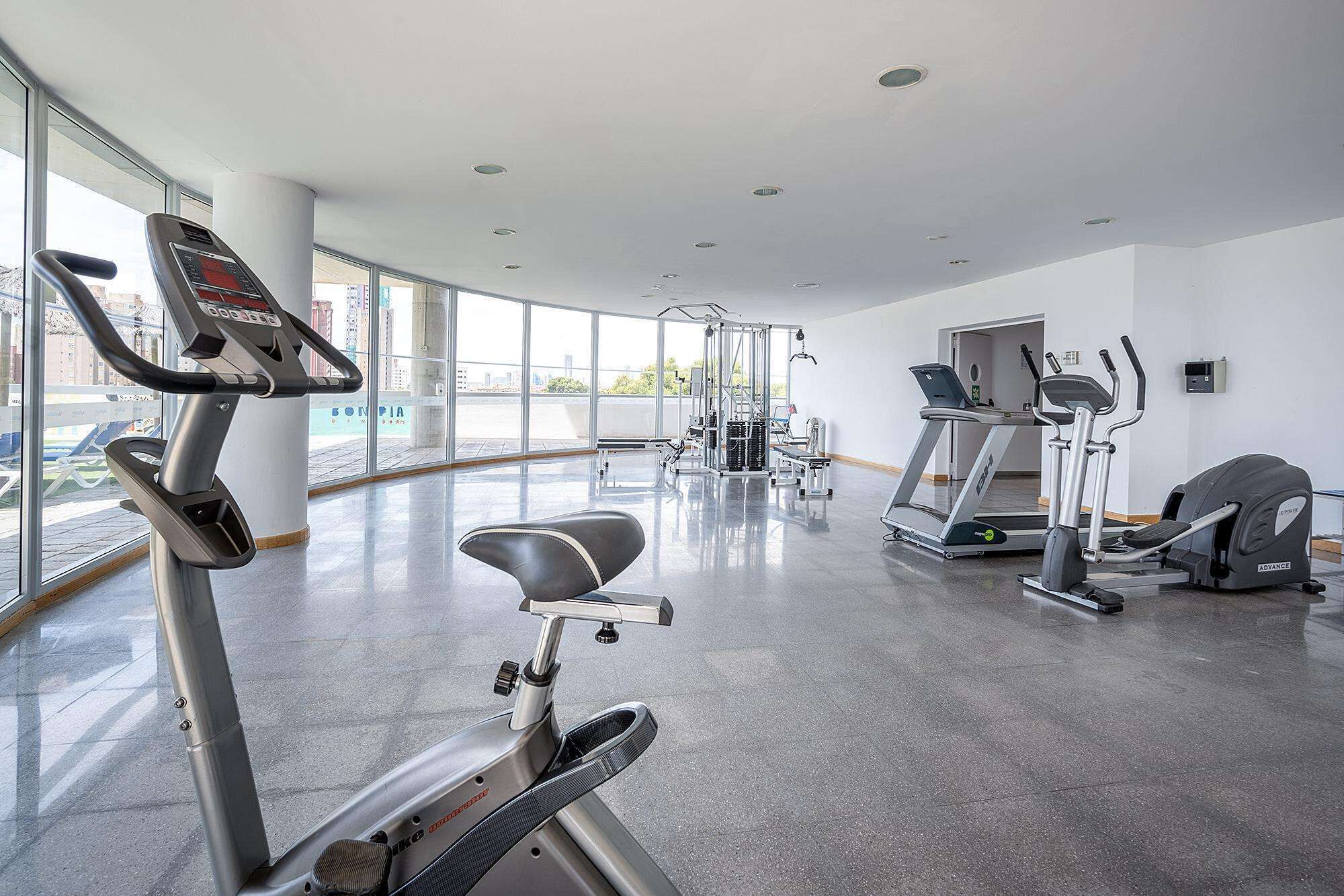 A modern gym with cardio machines, a weight station, and large windows offering views of the city.