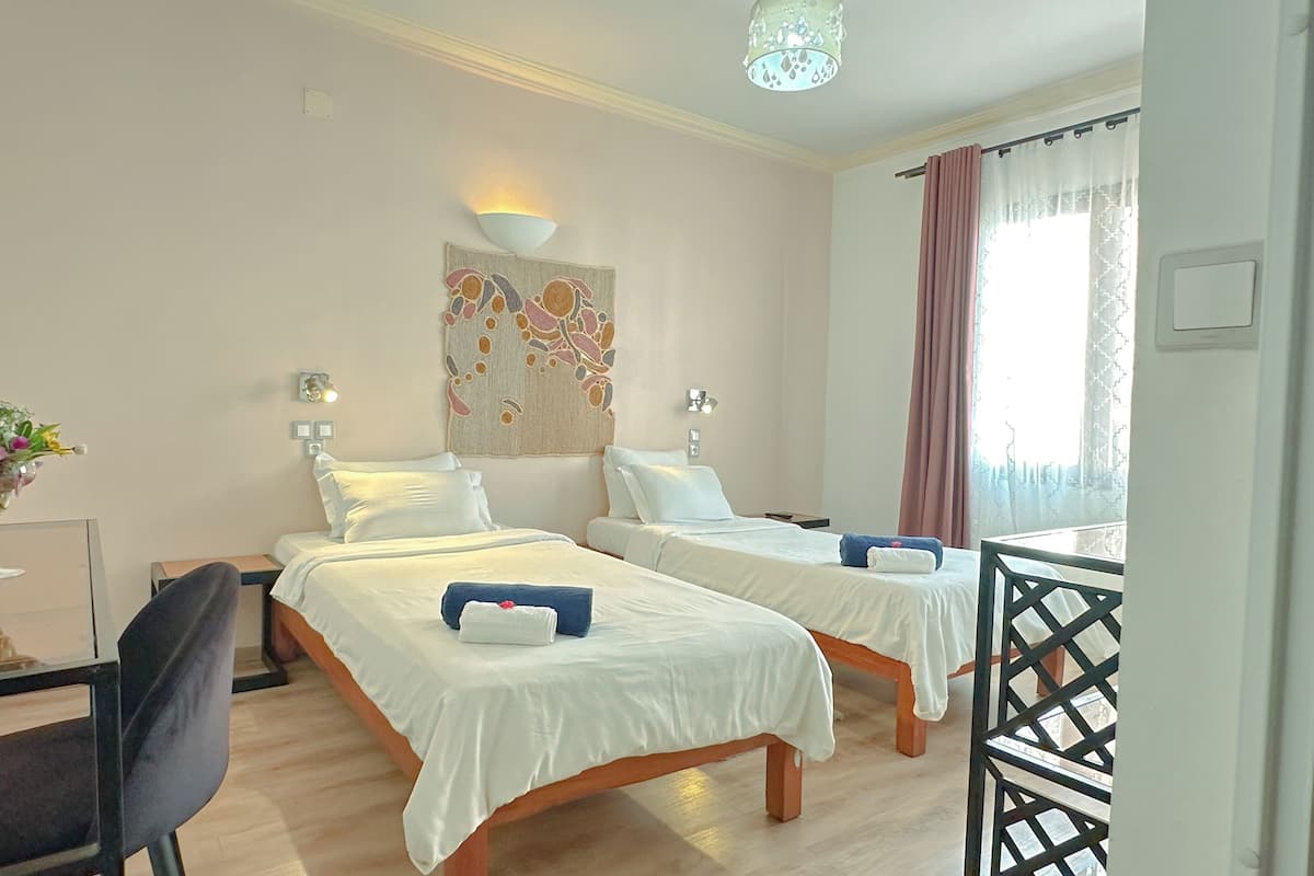 A hotel room with two single beds, a desk with a chair, a vase of flowers, and a decorative wall hanging.
