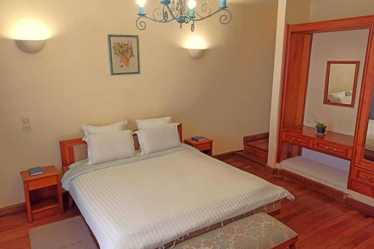 A bedroom with a large bed, bedside tables, a wooden bench, a chandelier, a wall-mounted mirror, and a framed picture.