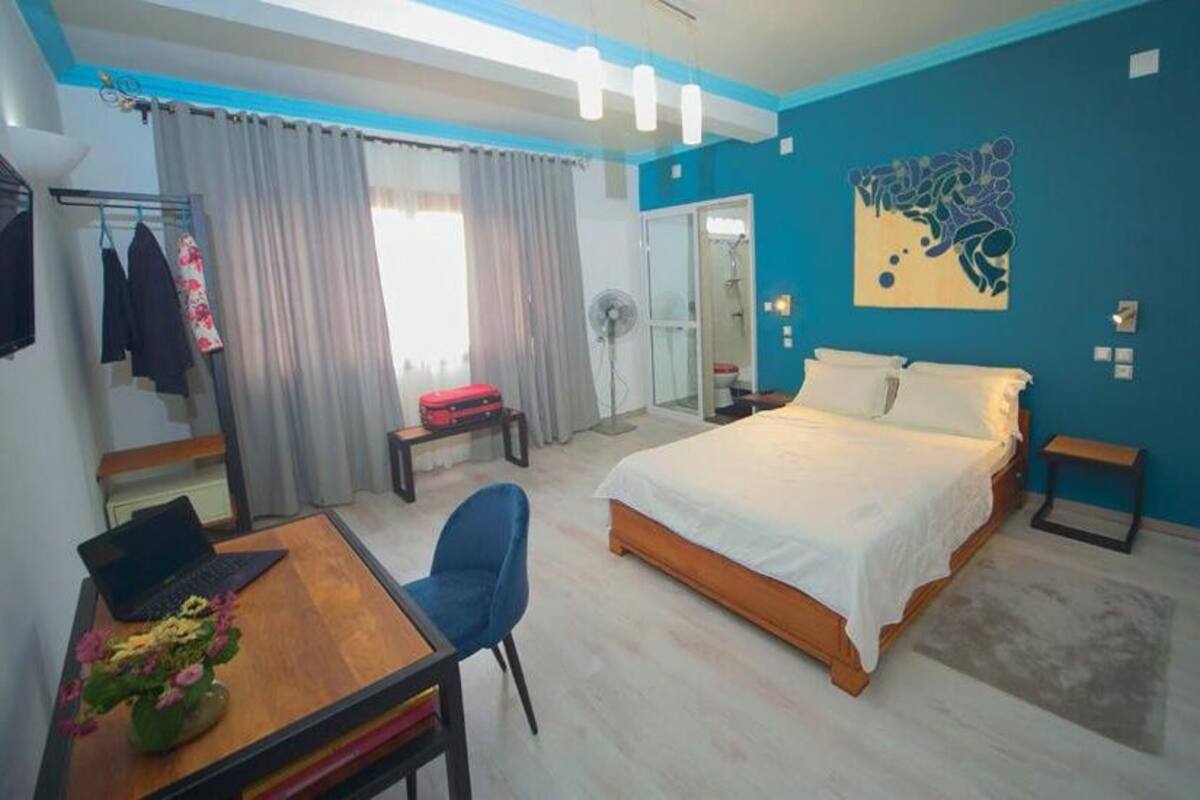 A modern bedroom with a bed, a desk with a laptop, a chair, a small table with flowers, a fan, and a sliding door leading to a bathroom.