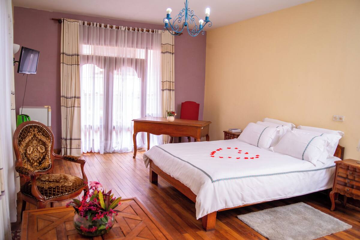 A bedroom with a bed, a desk, a chair, a television, and a chandelier.