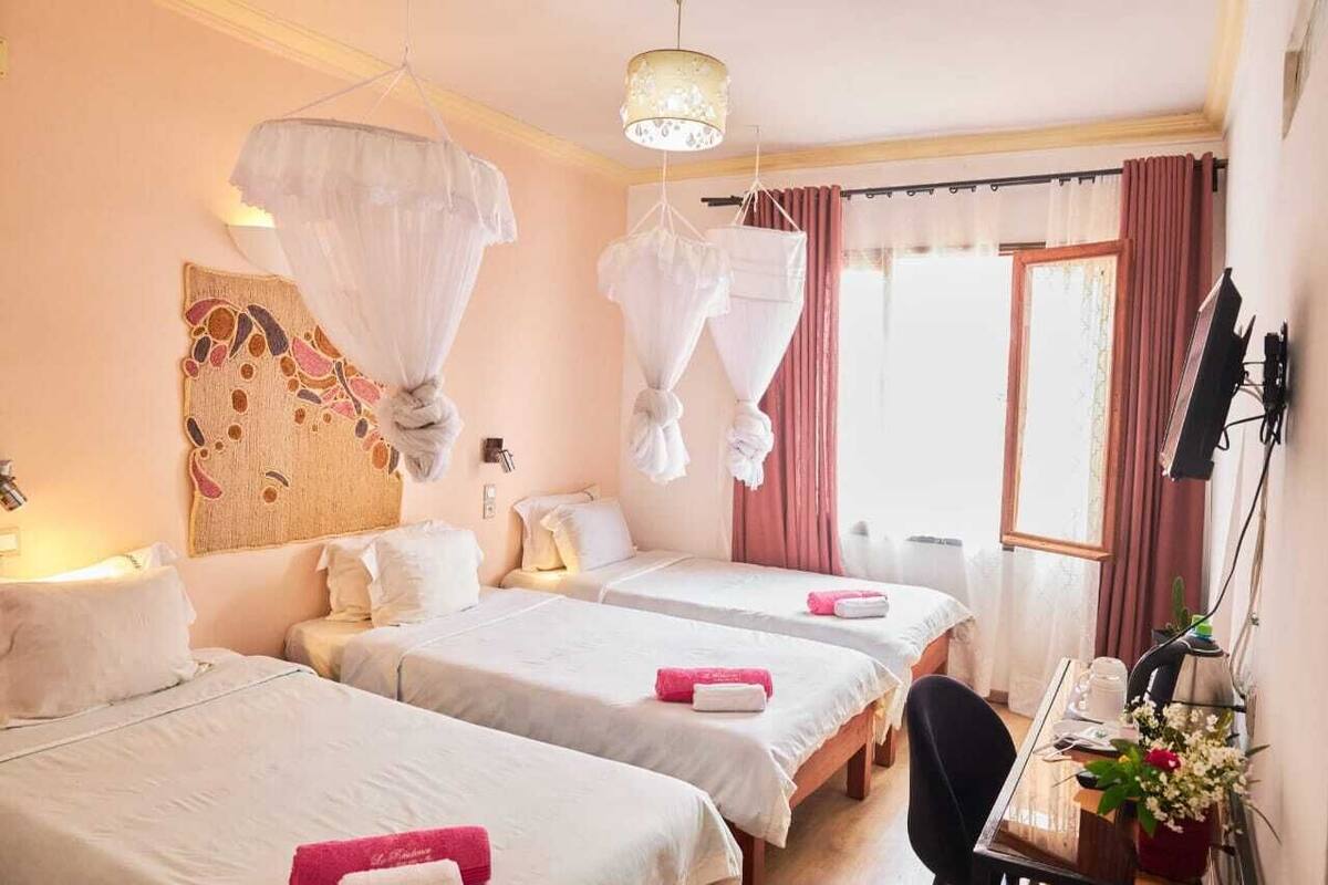 A hotel room with three beds, a desk, and a chair. There is a window with curtains, a wall-mounted artwork, and a hanging light fixture.