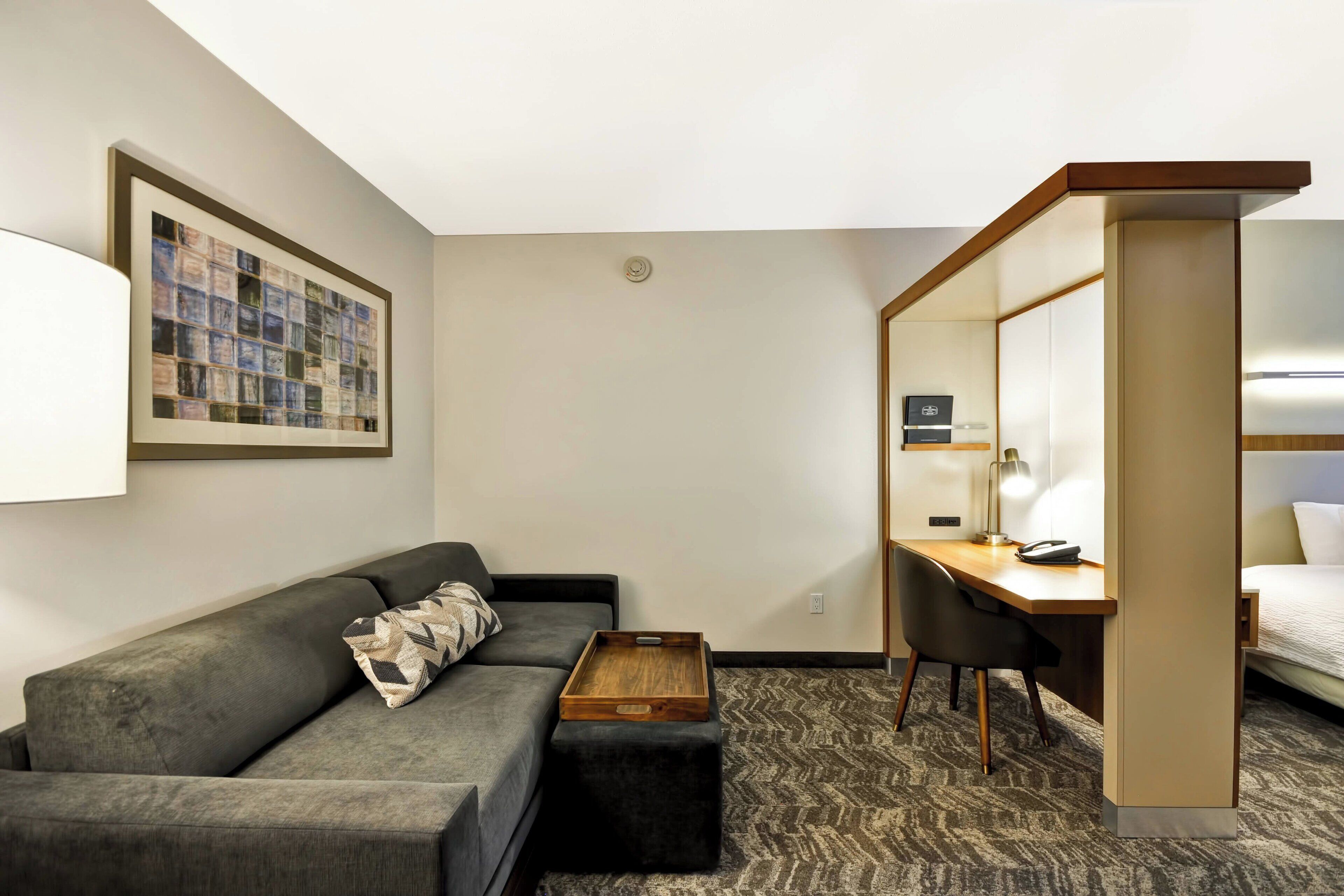 A hotel room with a gray sofa, a wooden side table, a desk with a chair, and a bed.