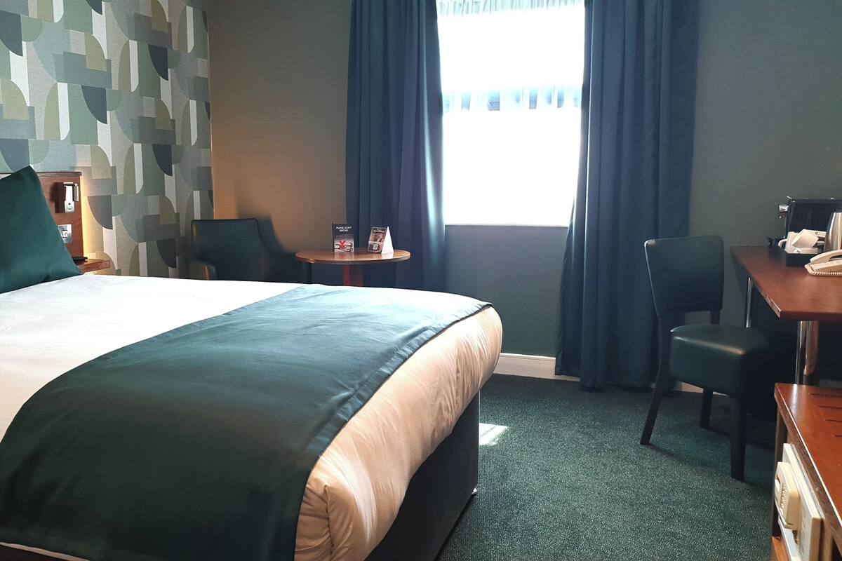 A hotel room with a bed, a desk, a chair, a small table, and a window with curtains.