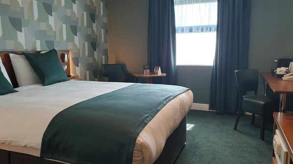 Habitación de hotel con una cama grande, un escritorio, una silla, una mesita y una ventana con cortinas.