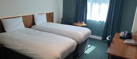 Une chambre d’hôtel avec deux lits, une petite table avec un téléphone, une chaise et une fenêtre avec des rideaux.