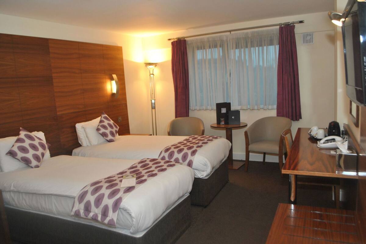 A hotel room with two beds, a desk, a chair, a lamp, and a window with curtains.