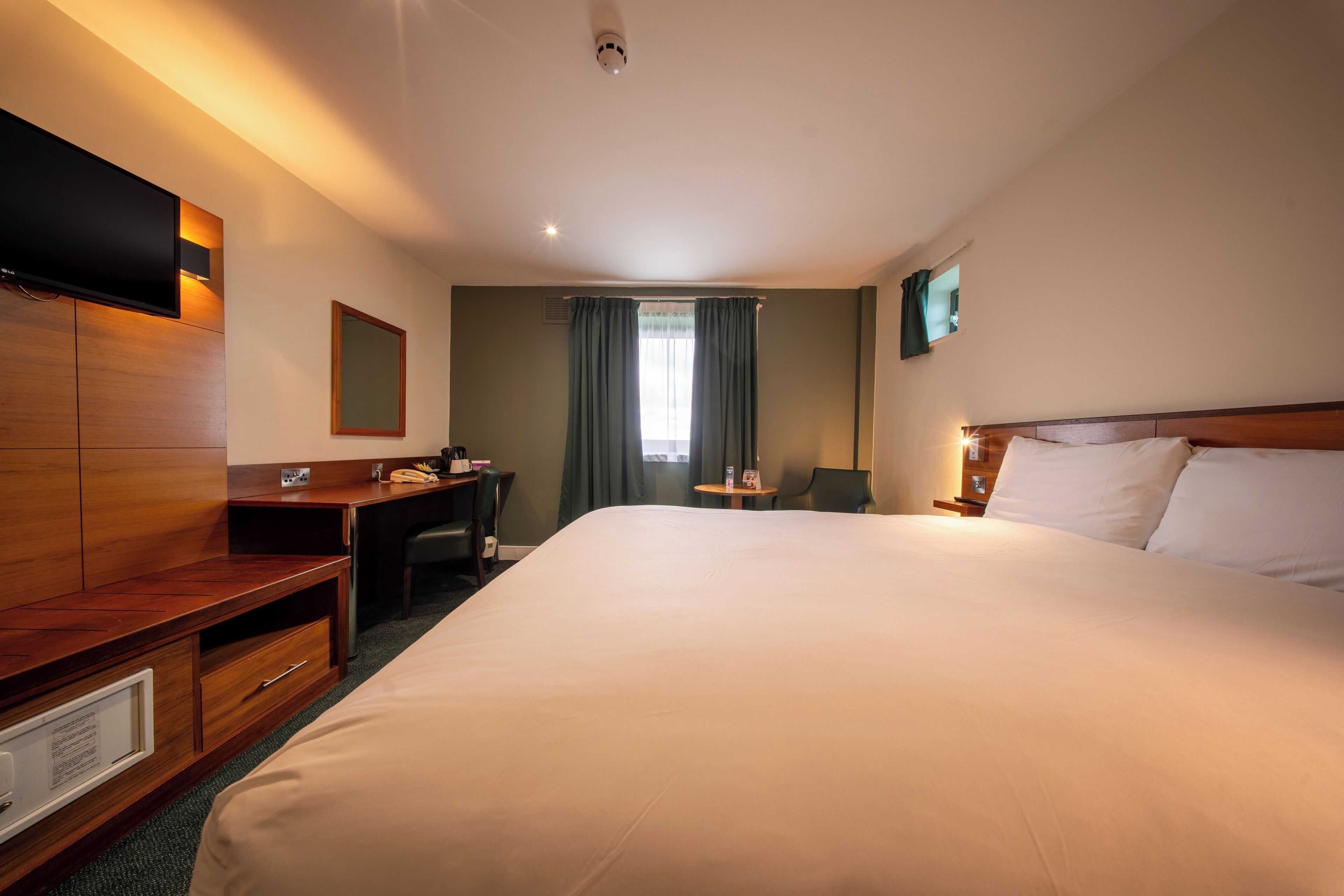 A hotel room with a large bed, a desk with a chair, a television mounted on the wall, and a window with curtains.