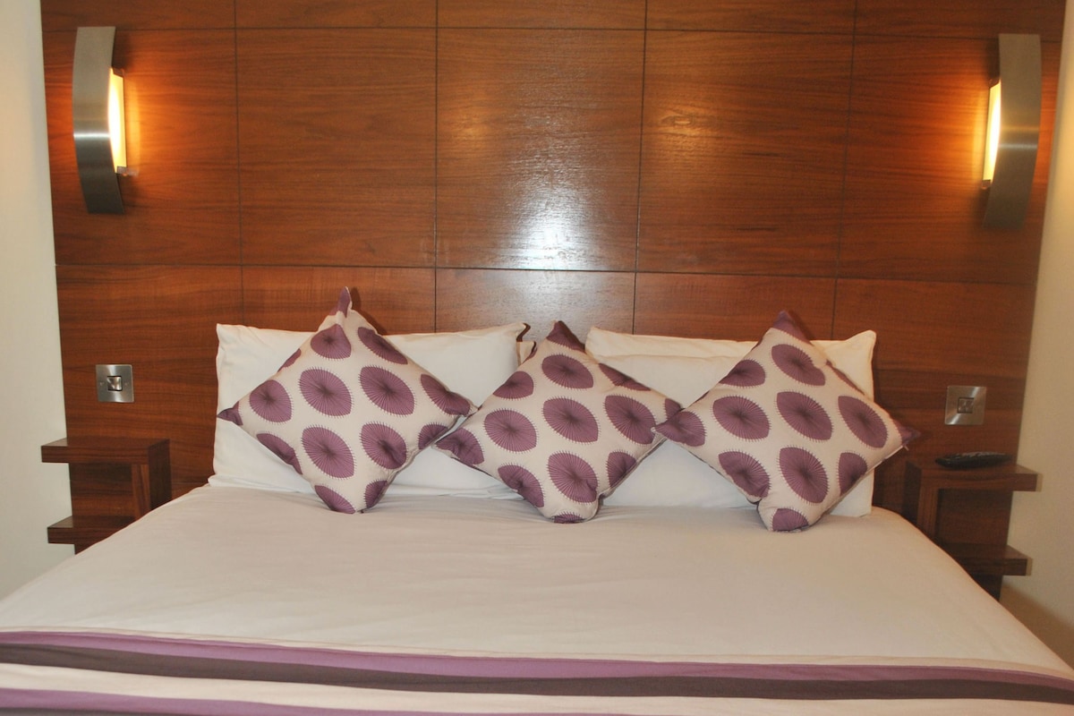 A hotel room with a wooden headboard, a bed with white bedding and purple patterned pillows, and two bedside lamps.