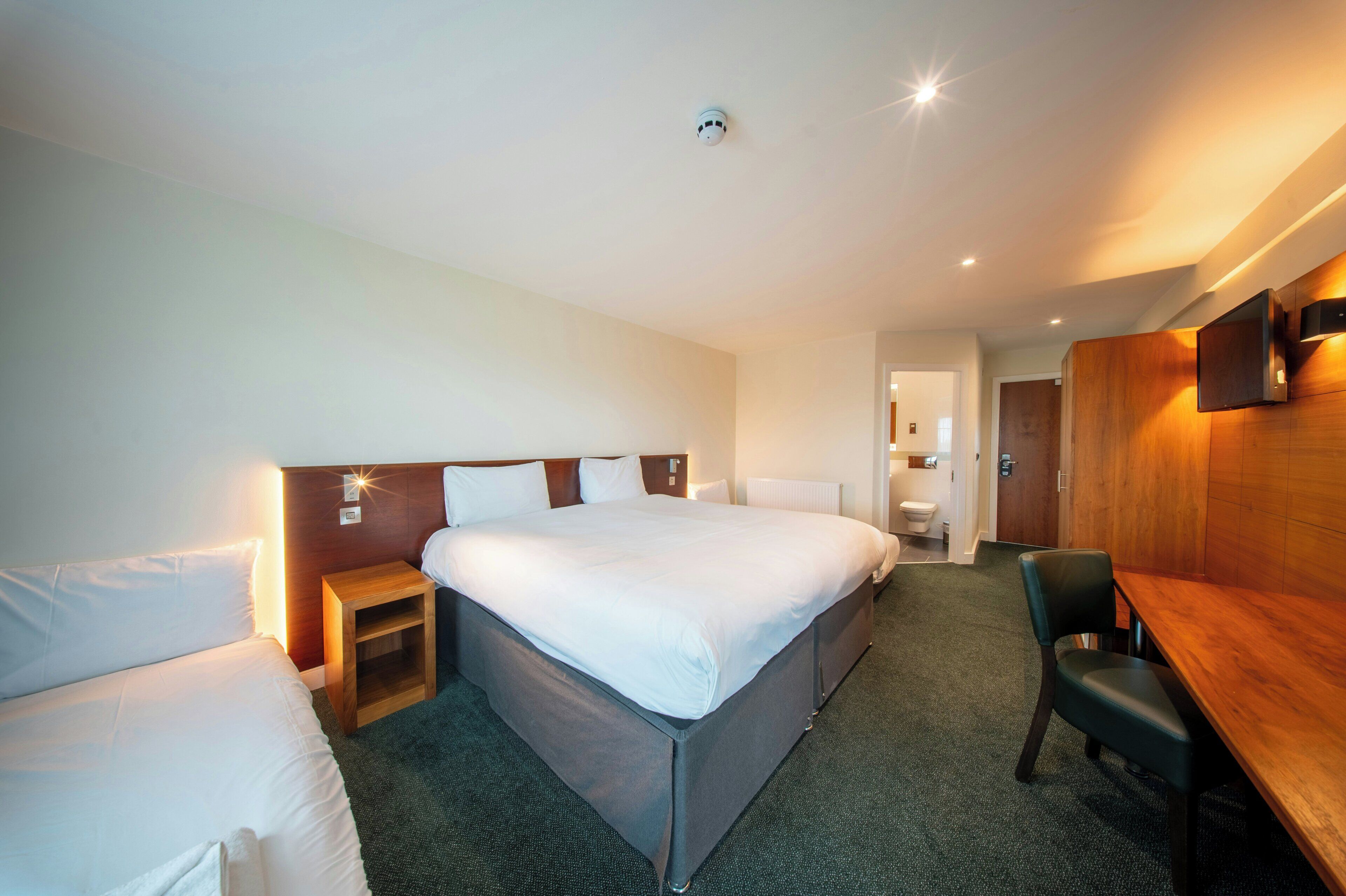 A hotel room with a large bed, a bedside table, a chair, a desk, and a bathroom visible through an open door.