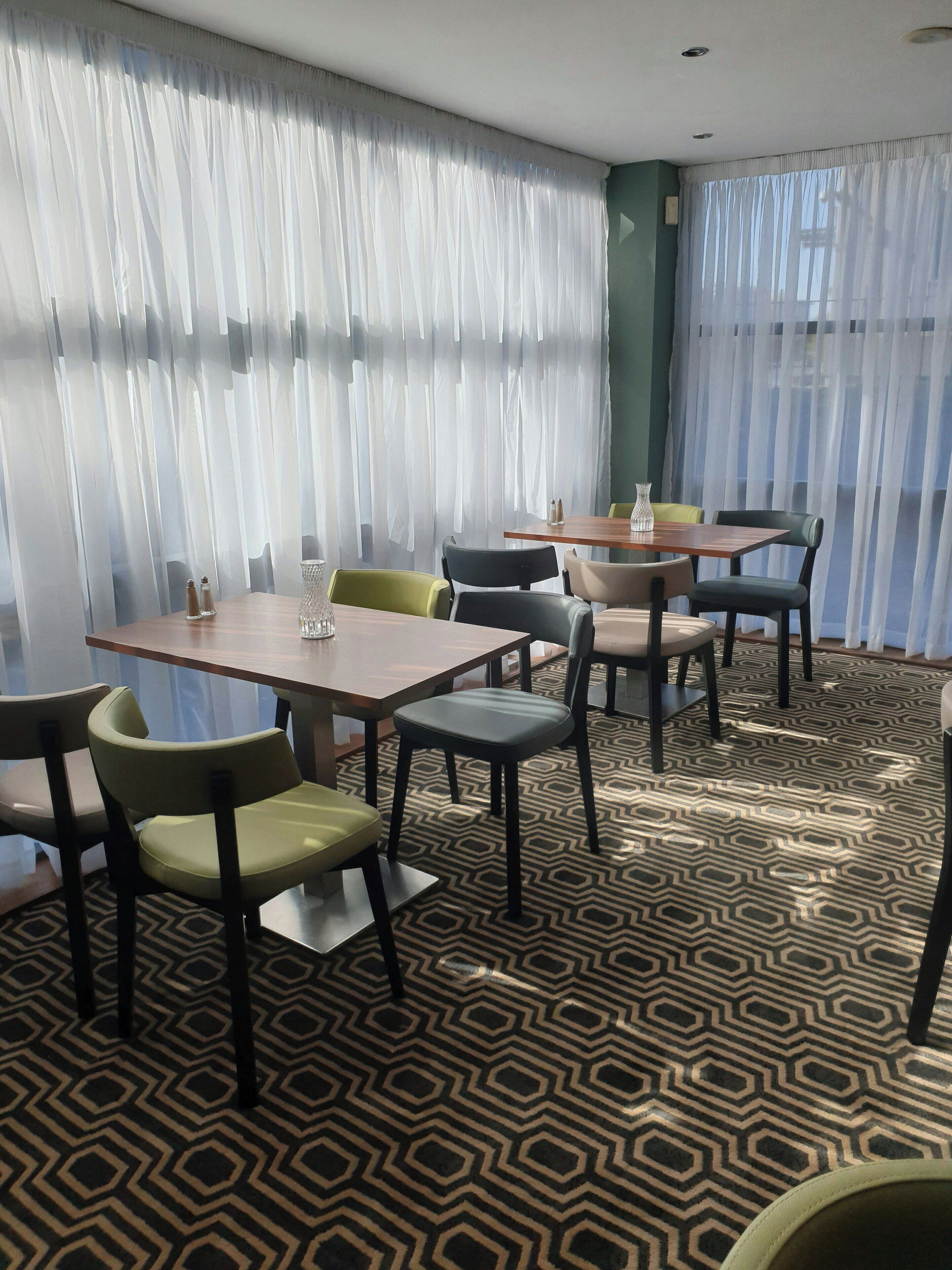 A hotel room with a dining area featuring tables and chairs, a patterned floor, and sheer curtains.