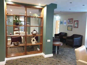 Eine Hotelhalle mit einem gemusterten Teppich, einem hölzernen Empfangsbereich, einer Sitzecke mit Stühlen und einer Wand mit gerahmten Kunstwerken.