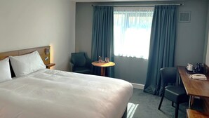 A hotel room with a bed, a desk, a chair, a small table, and a window with curtains.