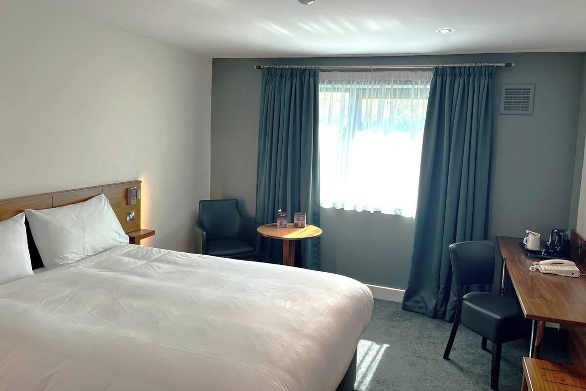 A hotel room with a bed, a desk, a chair, a small table, and a window with curtains.