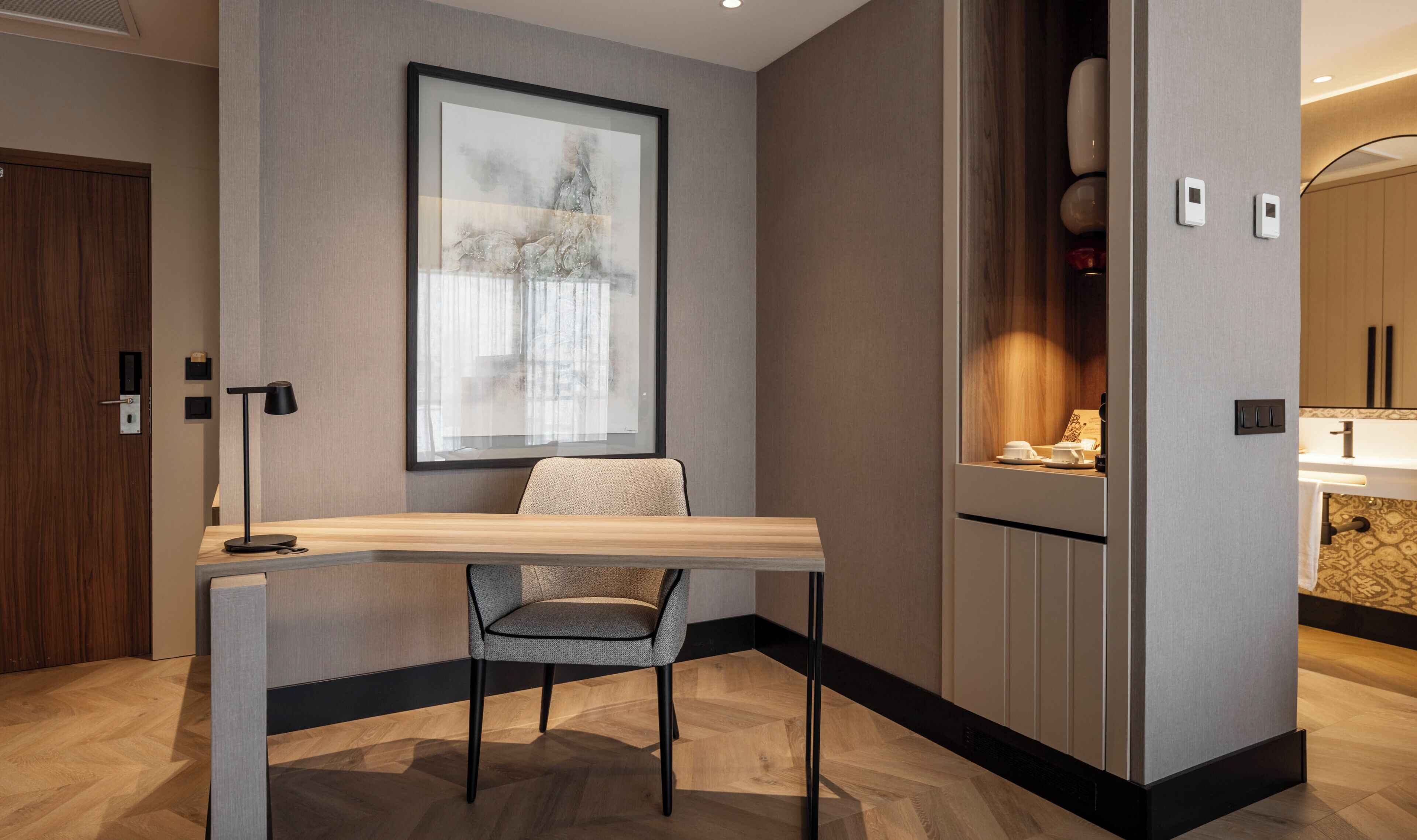 A modern hotel room with a wooden desk, a chair, a framed abstract painting, a window with curtains, and a bathroom visible through a door.