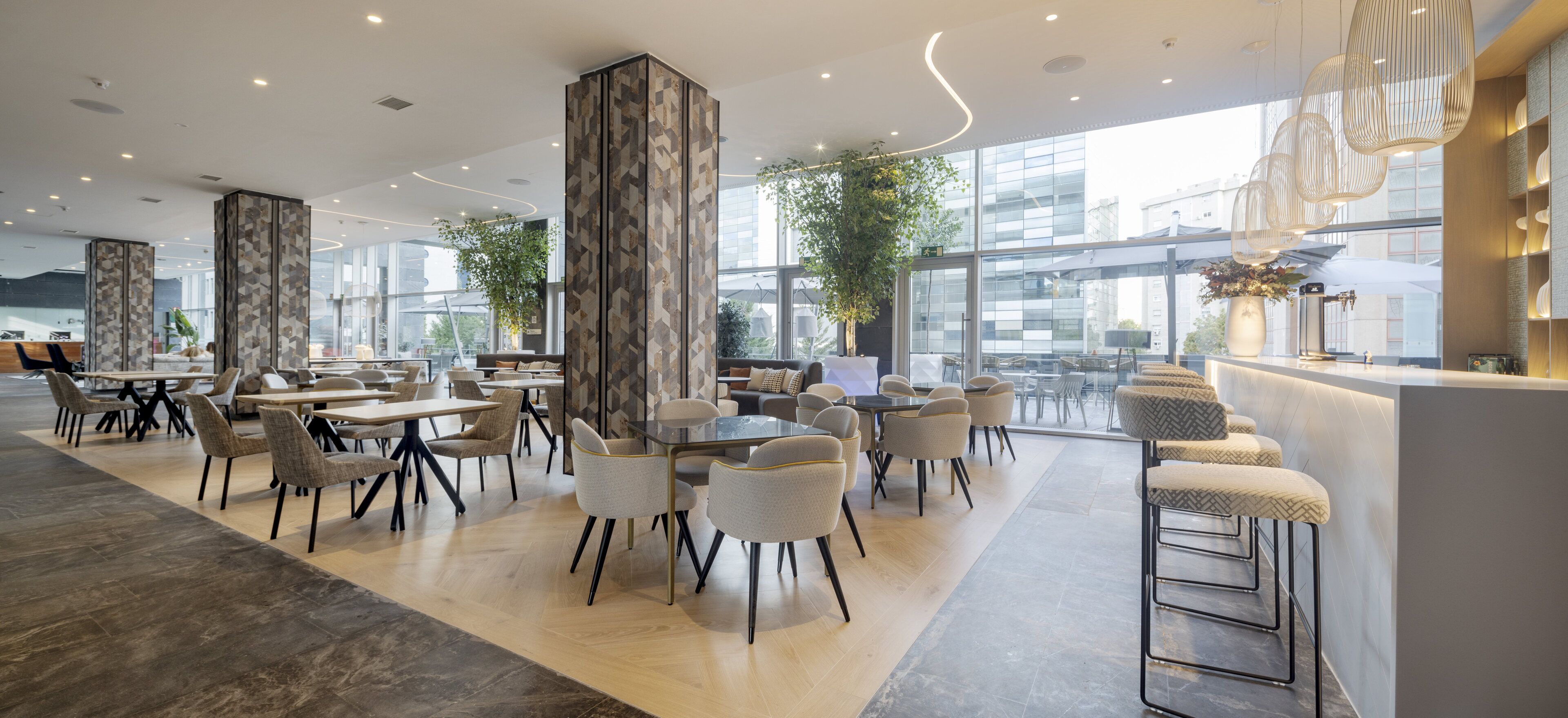 Un moderno vestíbulo de hotel con una variedad de espacios de descanso, incluyendo mesas y sillas, una zona de bar y amplios ventanales con vistas a la ciudad.