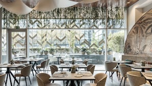 Un restaurant avec des tables et des chaises, de grandes fenêtres donnant sur un espace verdoyant et des plantes suspendues.