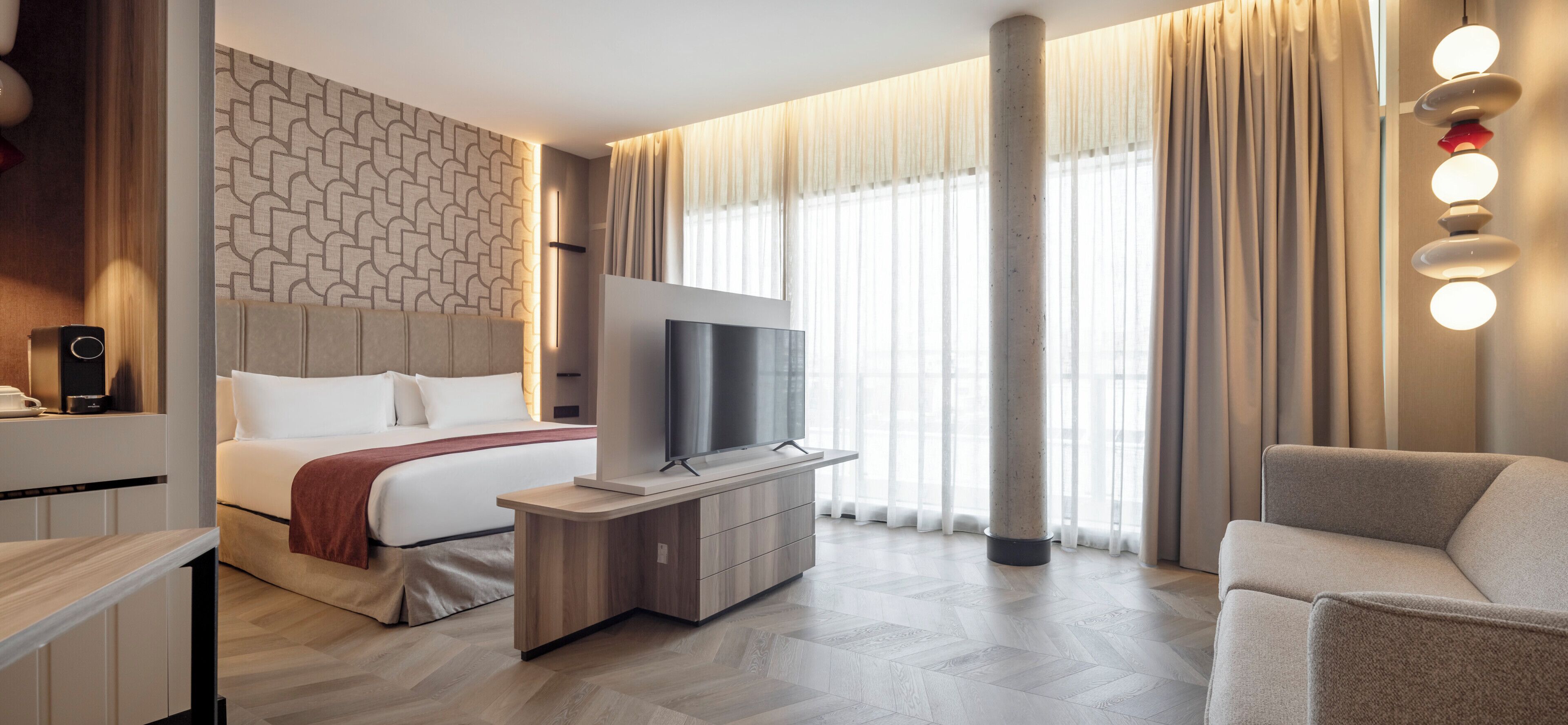 A modern hotel room with a bed, TV, seating area, and large windows.