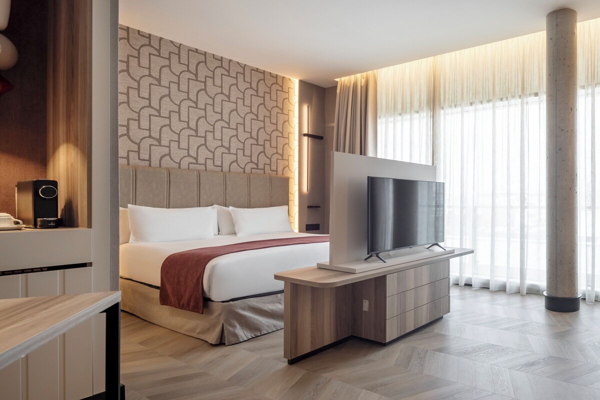 Quarto de hotel moderno com cama, televisão, área de estar e grandes janelas.