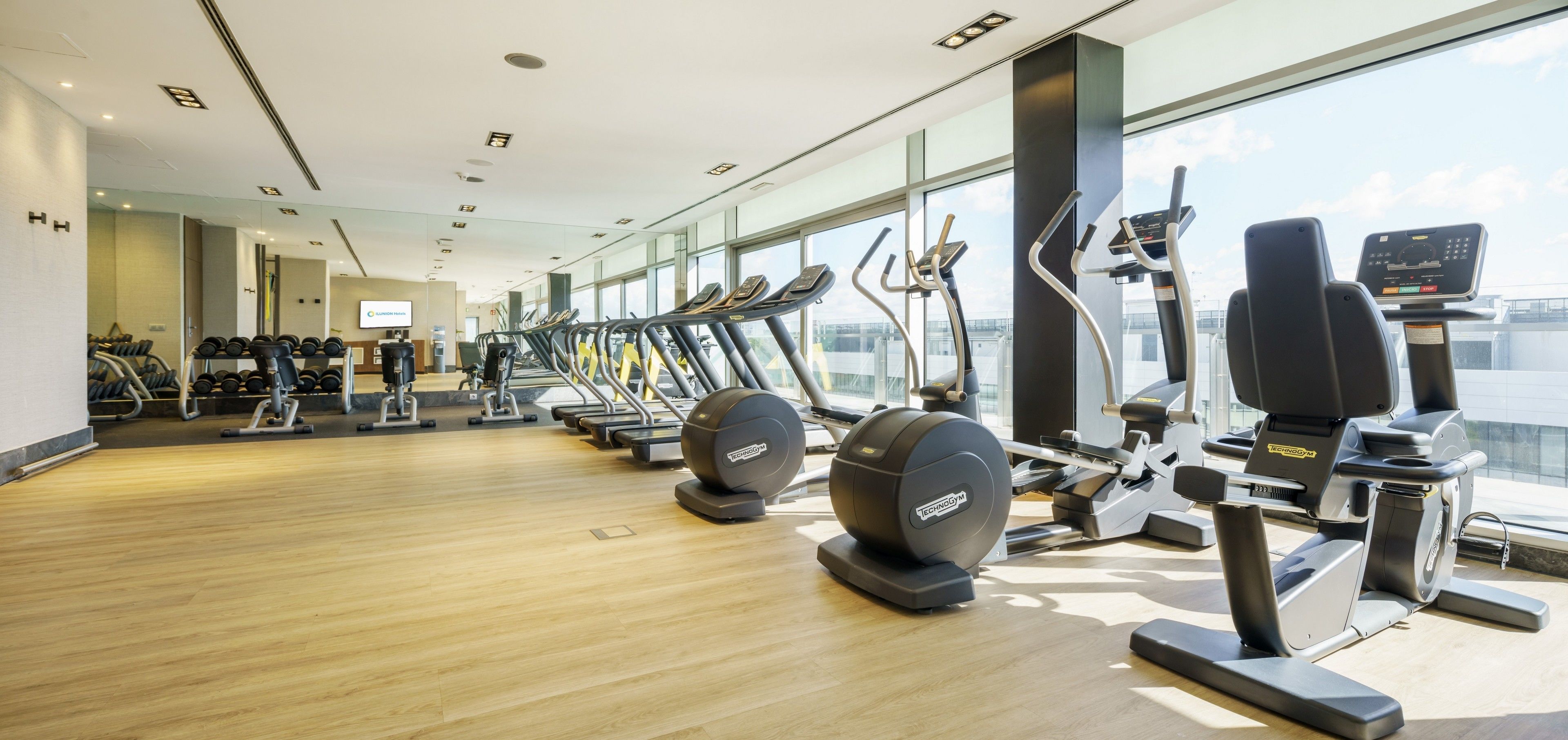 A modern gym with various cardio machines, a wooden floor, and large windows offering a city view.