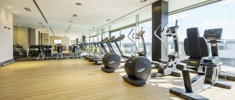 A modern gym with various cardio machines, a wooden floor, and large windows offering a city view.