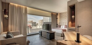 A modern hotel room with a large window, a flat-screen TV, a bed, a sofa, and a desk.