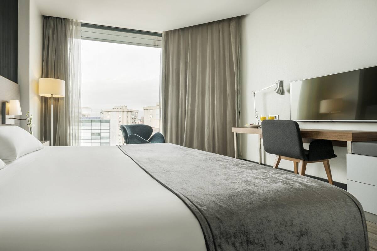 Una camera d'albergo con un letto grande, una scrivania con una sedia, una TV e vista sulla città dalla finestra.
