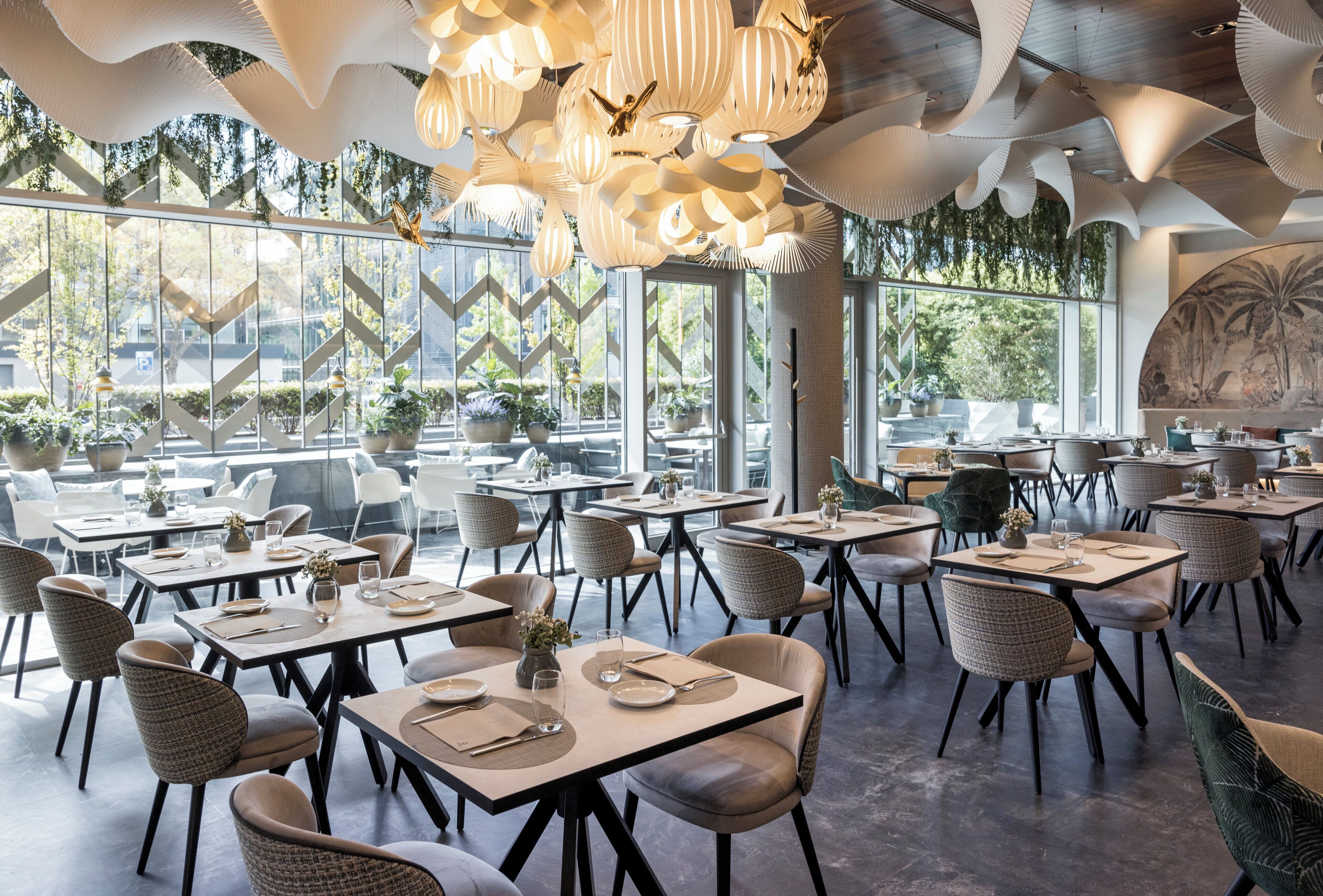 A modern restaurant with high ceilings, unique lighting fixtures, and large windows offering views of greenery.
