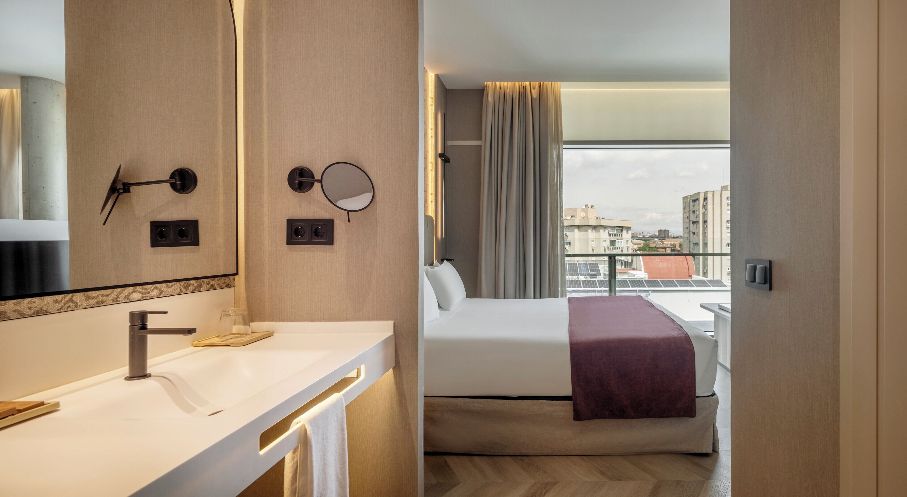 A hotel room with a modern bathroom, a bed with a view, and a cityscape visible through the window.