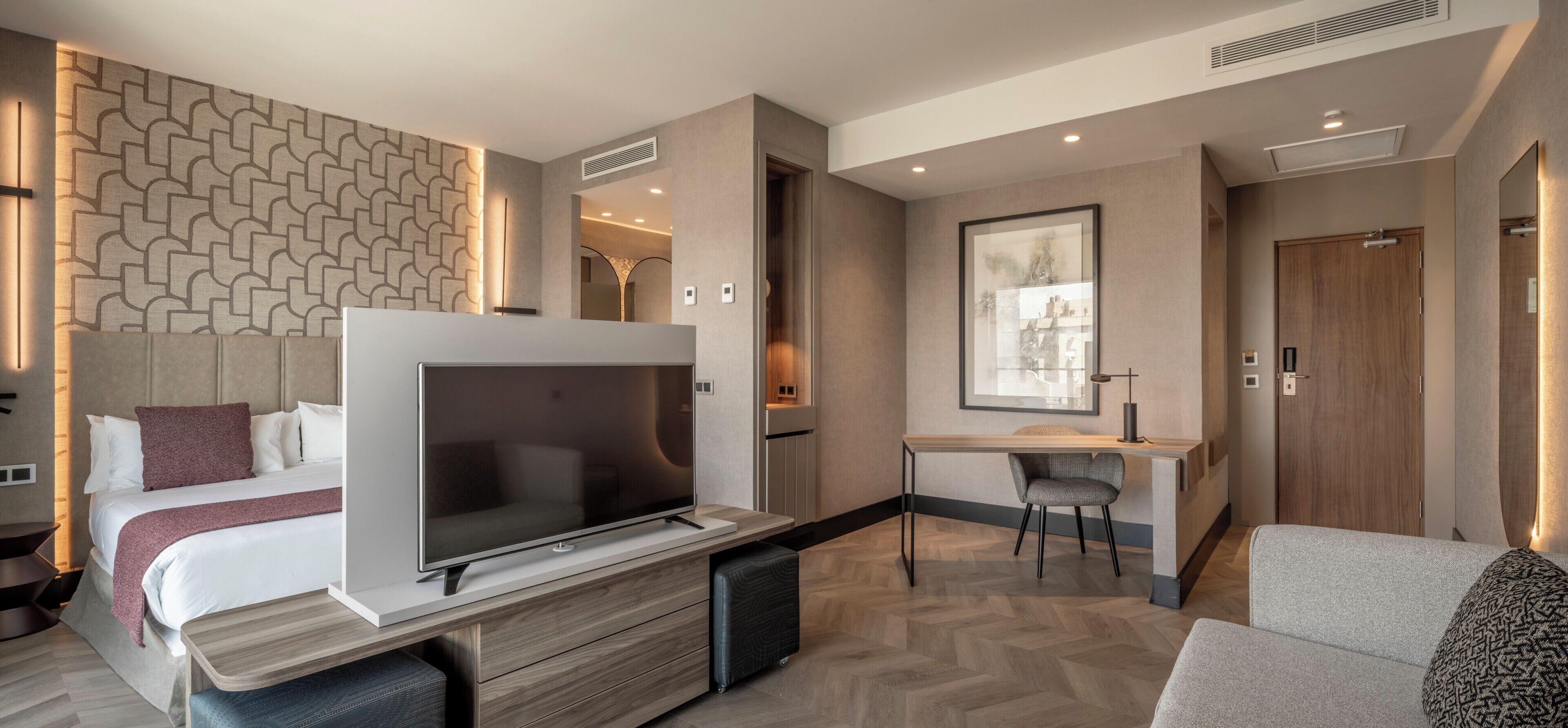 A modern hotel room with a large flat-screen TV, a bed with a headboard, a desk with a chair, and a large window.