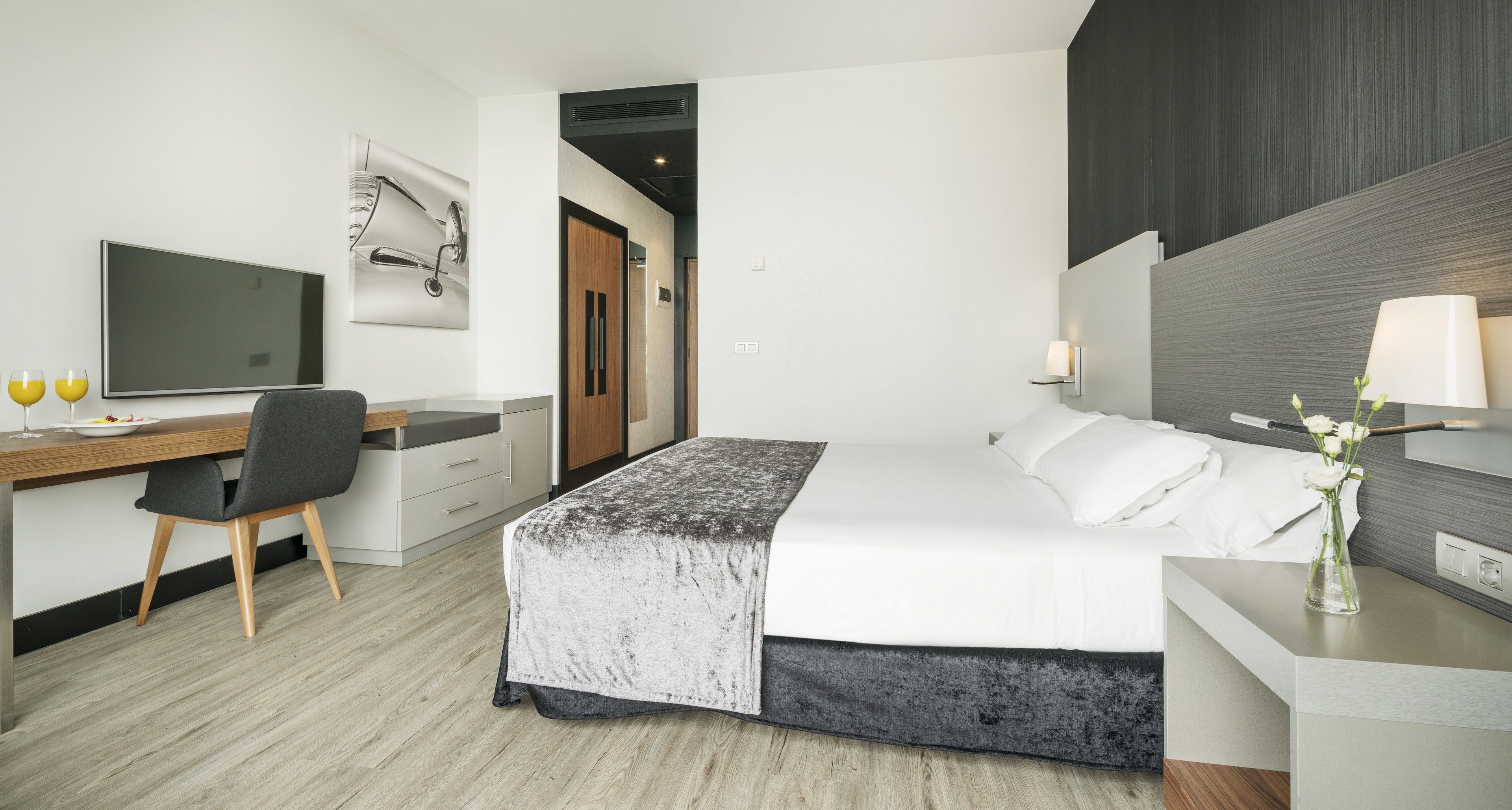 Una habitación de hotel moderna con cama, escritorio, silla, televisor y mesita de noche con un florero.