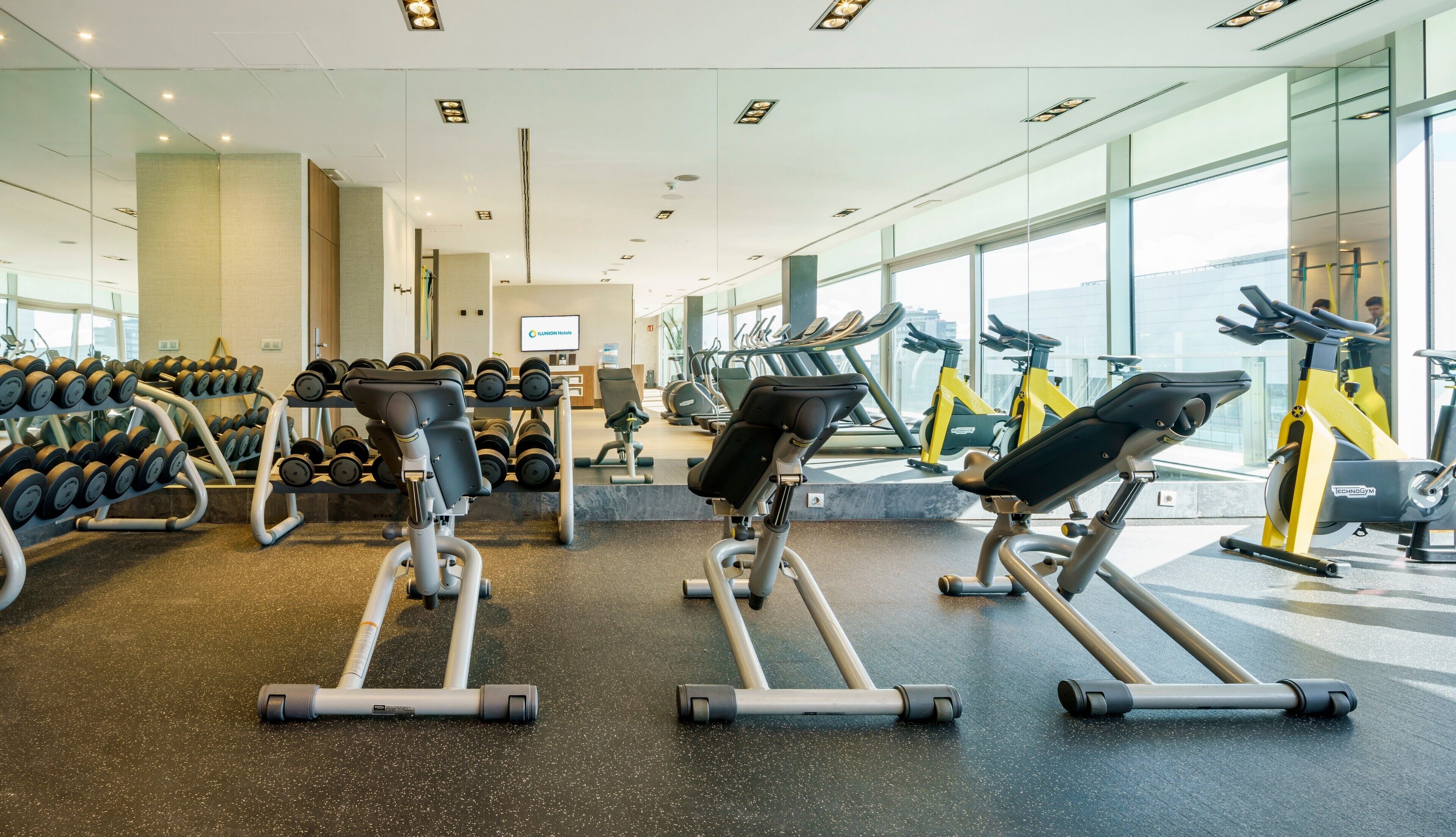 Une salle de sport équipée de machines d’exercice, de supports pour les poids et dotée de grandes fenêtres.