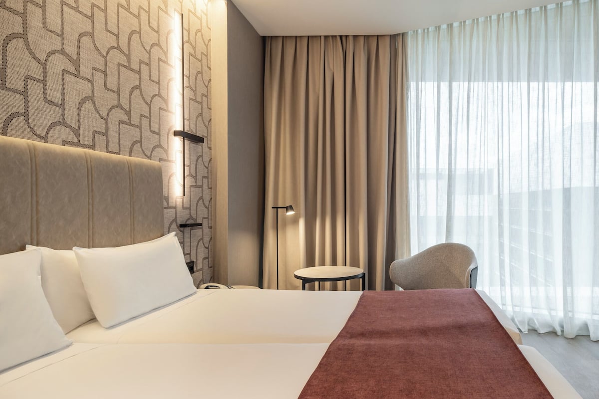 Una camera d'albergo con un letto grande, un comodino, una sedia, una panca e un'ampia finestra con tende.