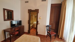 A room with a television on a wooden desk, a chair, a bathroom visible through an open door, and a large wooden door with curtains. - Hotel Coso Viejo (Antequera)
