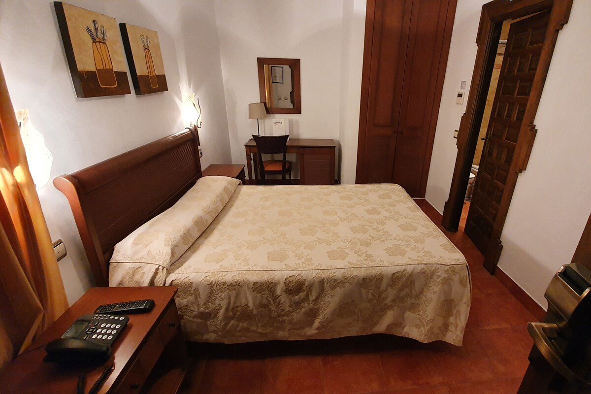 A bedroom with a wooden bed, a bedside table with a telephone, a desk with a lamp, and a door.