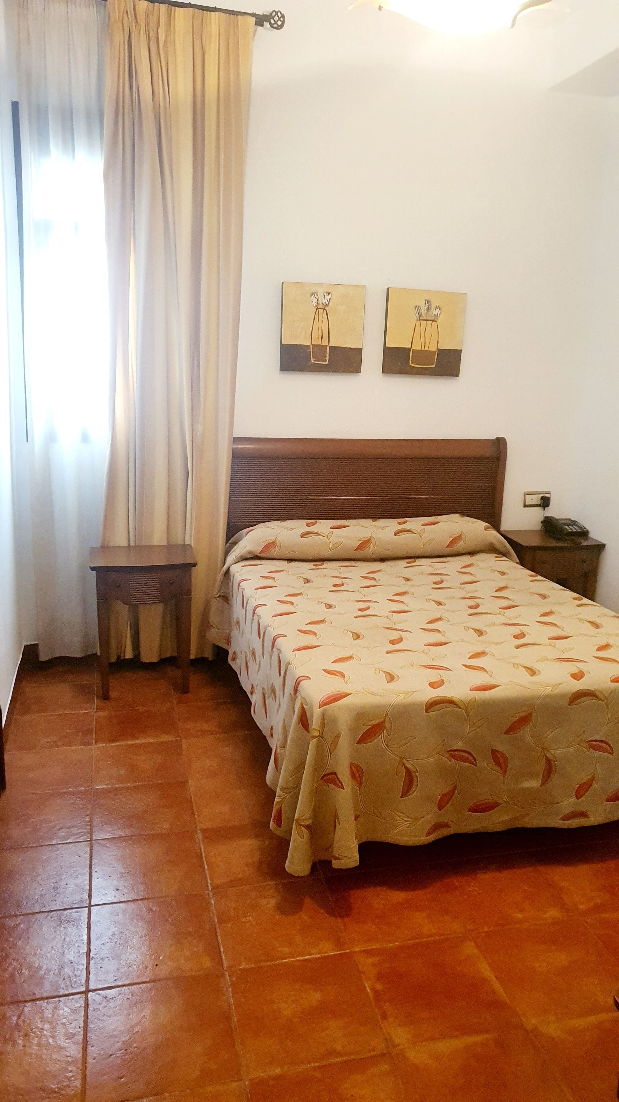 Un dormitorio con cama, mesita de noche, una pequeña mesa y dos cuadros colgados en la pared.