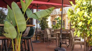 An outdoor patio with terracotta tiles, wicker furniture, and potted plants under red umbrellas.