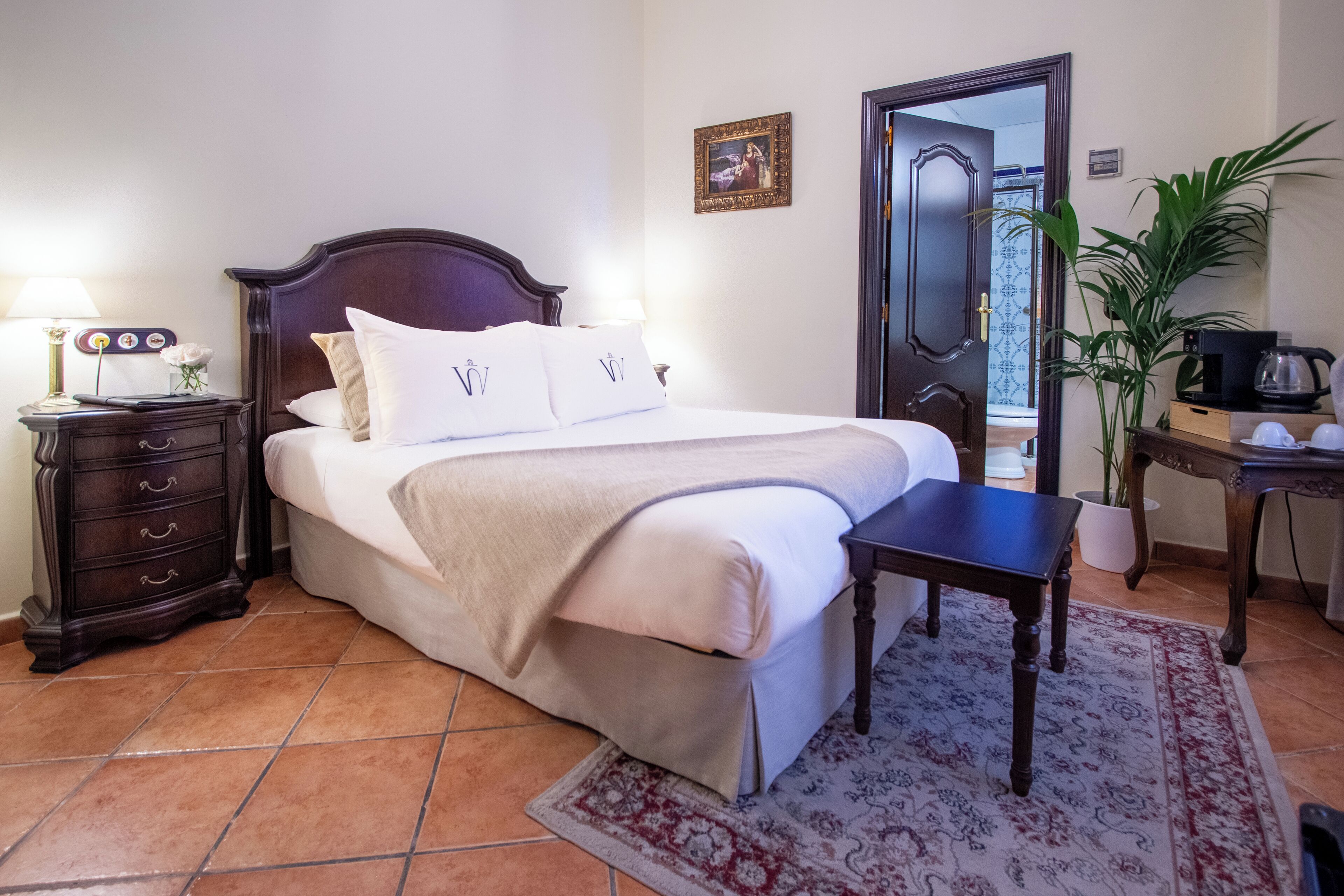 A neatly made bed with a headboard, a bedside table with a lamp, a small plant, and a coffee maker on a desk.