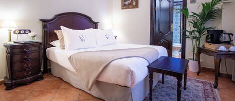 A neatly made bed with a headboard, a bedside table with a lamp, a small plant, and a coffee maker on a desk.