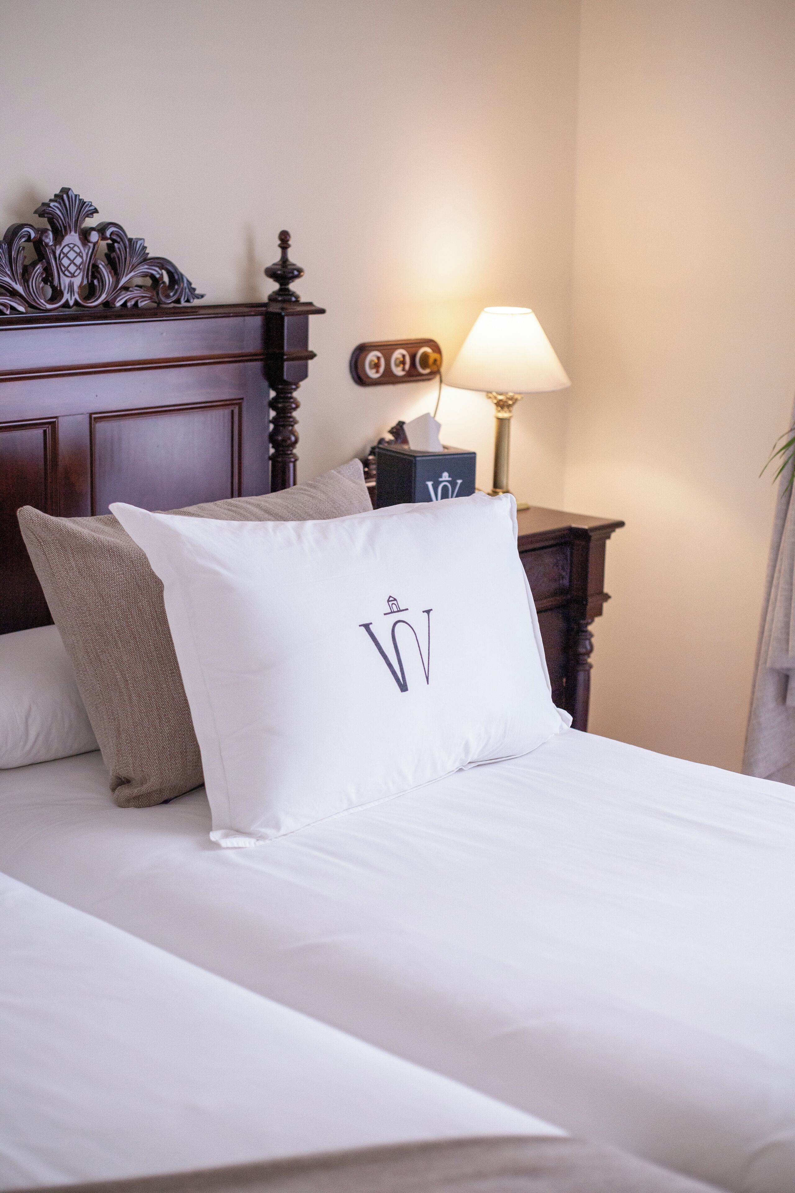 Una cama bien hecha con sábanas blancas y almohadones decorativos, un cabecero de madera, una mesita de noche con una lámpara y un reloj de pared.