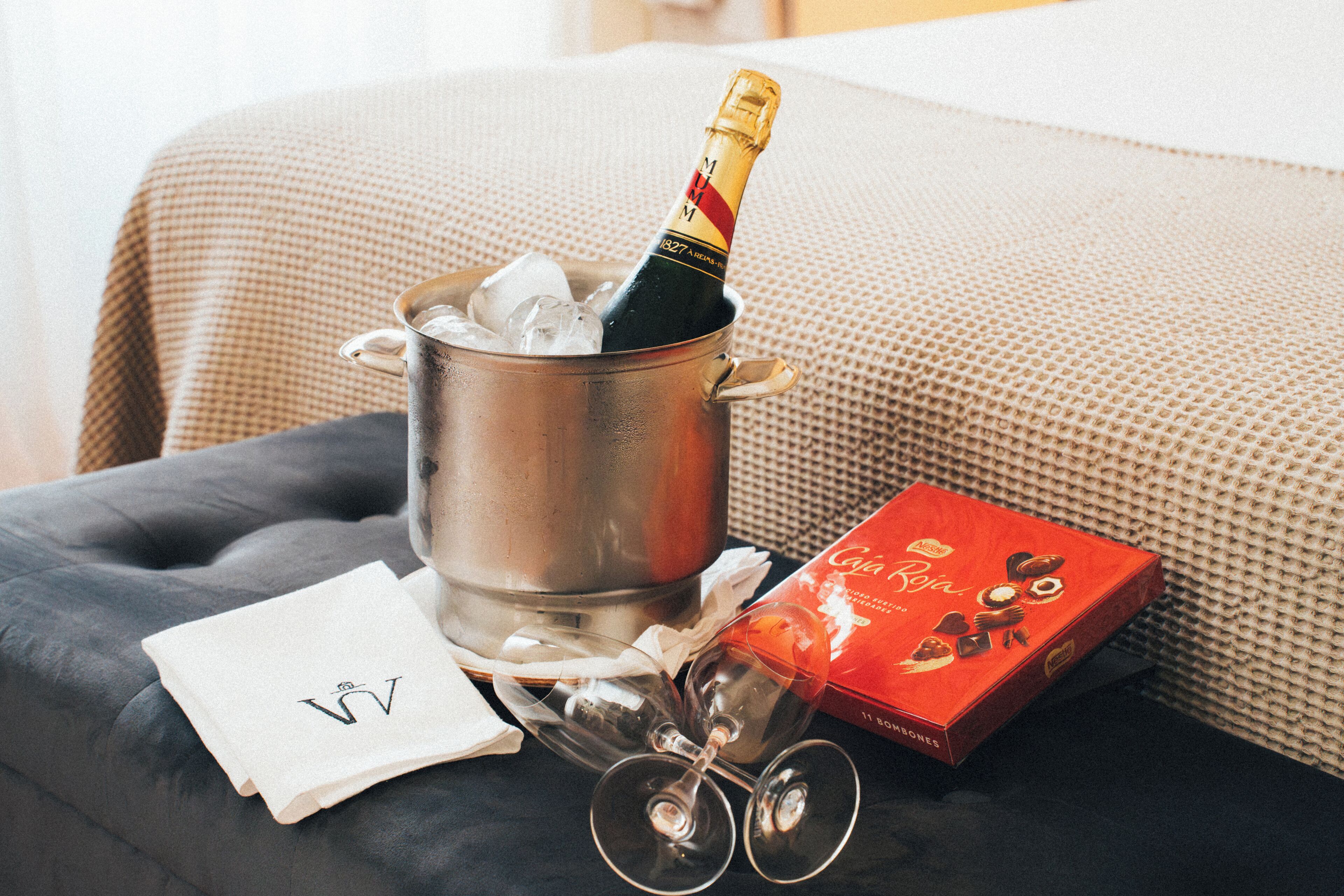 A metal bucket with a bottle of champagne, a box of chocolates, and two wine glasses on a dark surface.