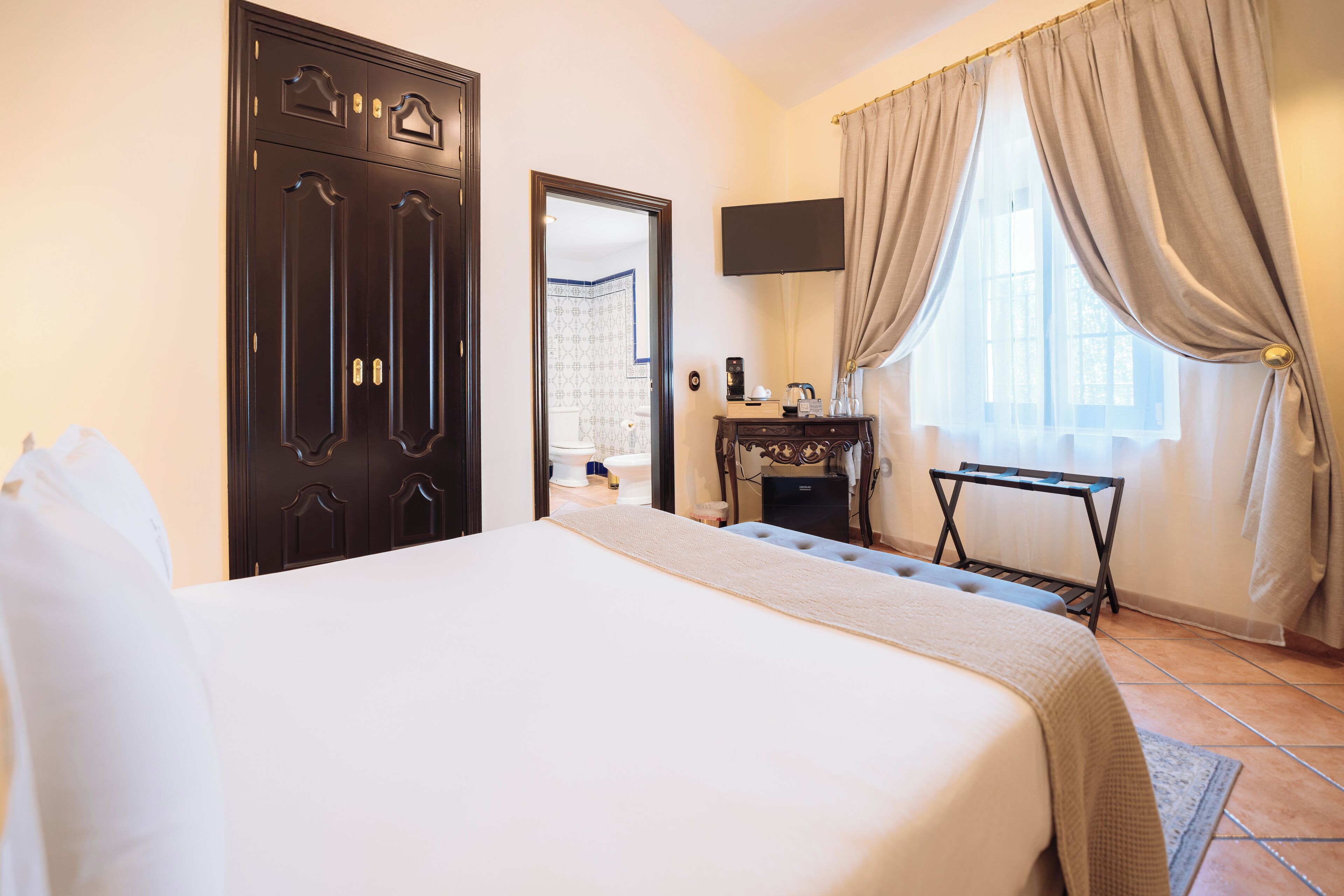 A hotel room with a large bed, a wardrobe, a desk with a flat-screen TV, and a window with curtains.