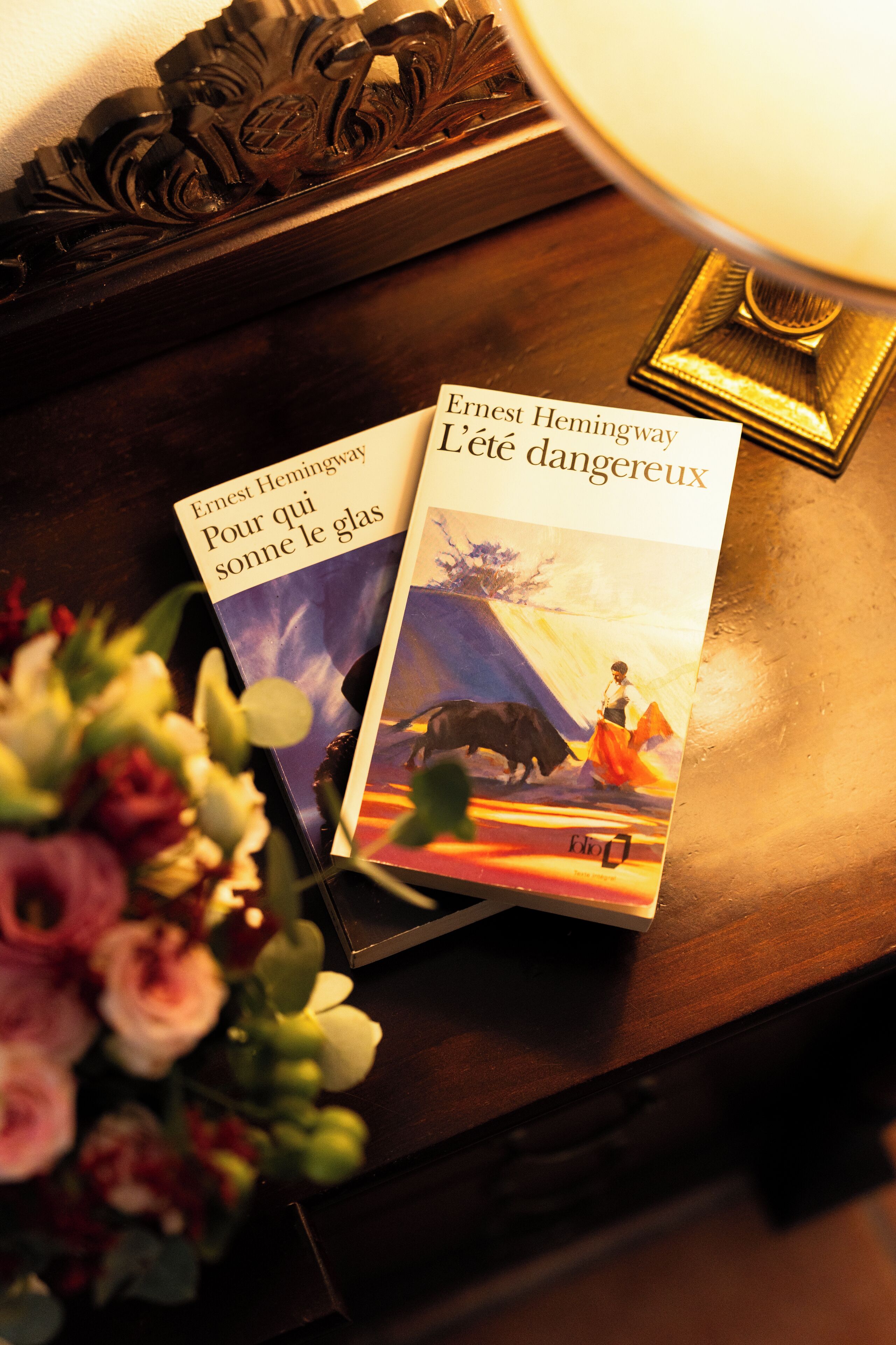 Two books titled 'Pour qui sonne le glas' and 'L'éte dangereux' by Ernest Hemingway on a wooden surface with a lamp and a bouquet of flowers.