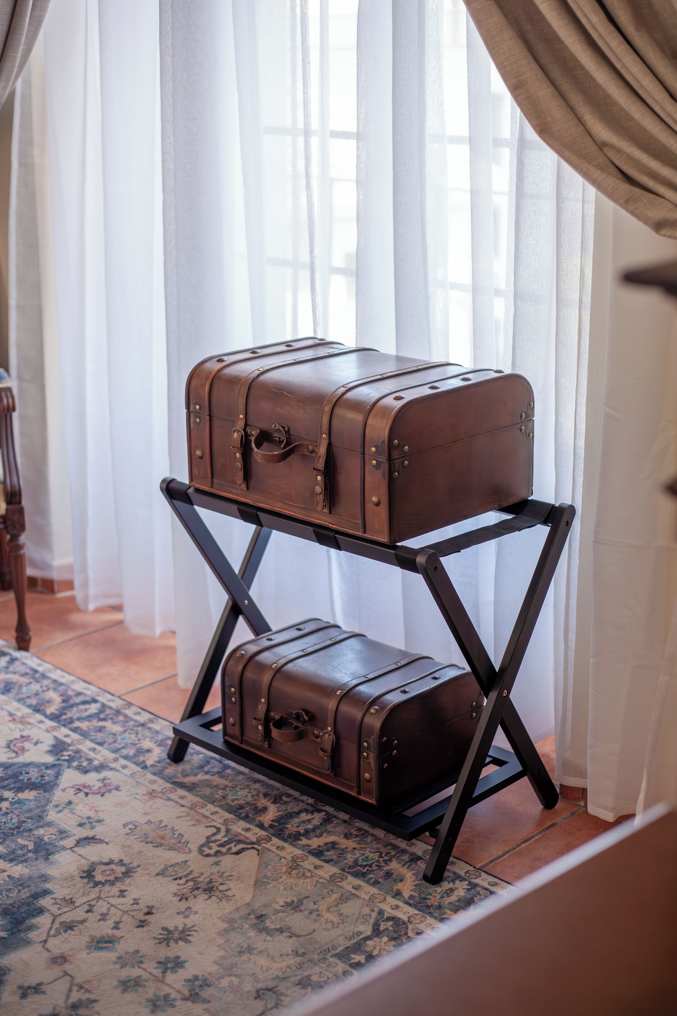 A room with a vintage suitcase on a stand near a window with sheer curtains.
