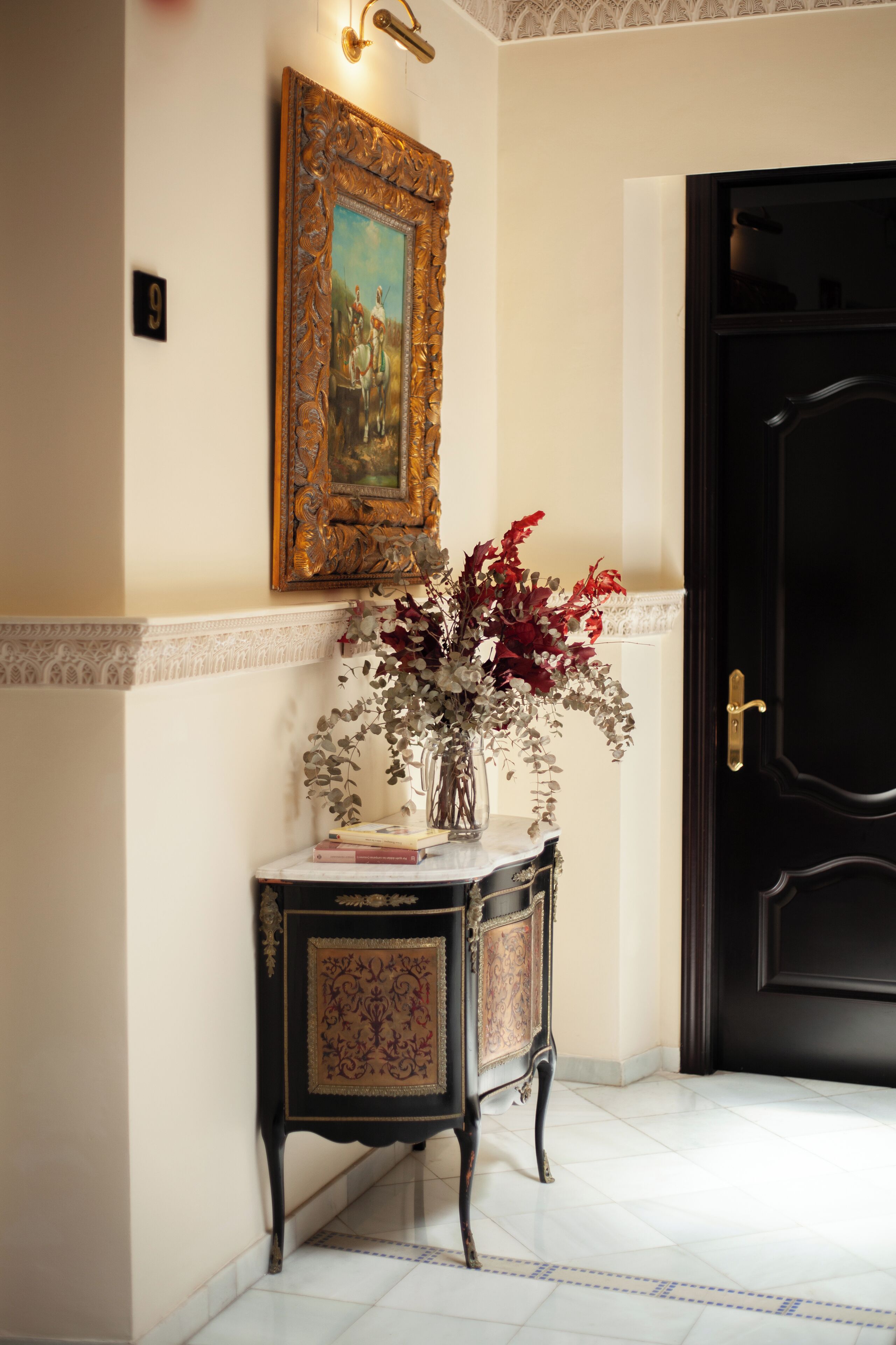 A hallway with a black door, a framed painting on the wall, and a small table with a vase of flowers and books.
